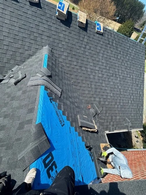 Worker installing a new shingle roof with black shingles, blue underlayment, and ventilation pipes on a residential house.