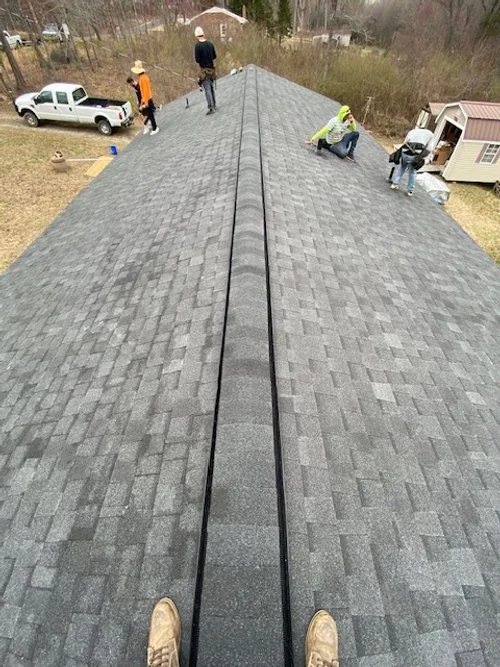 View from the top of a house roof with four workers installing or inspecting the shingles, and a person taking the photo from the edge.