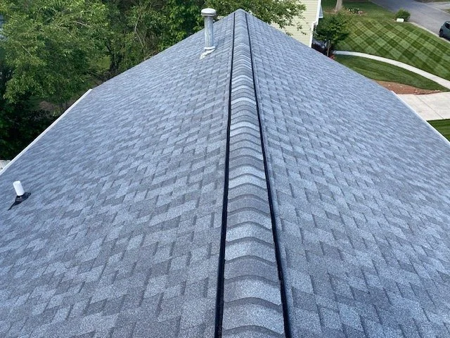 View of a house roof with asphalt shingles, ridge vent in the center, and vent pipes on either side, with trees and a street in the background.