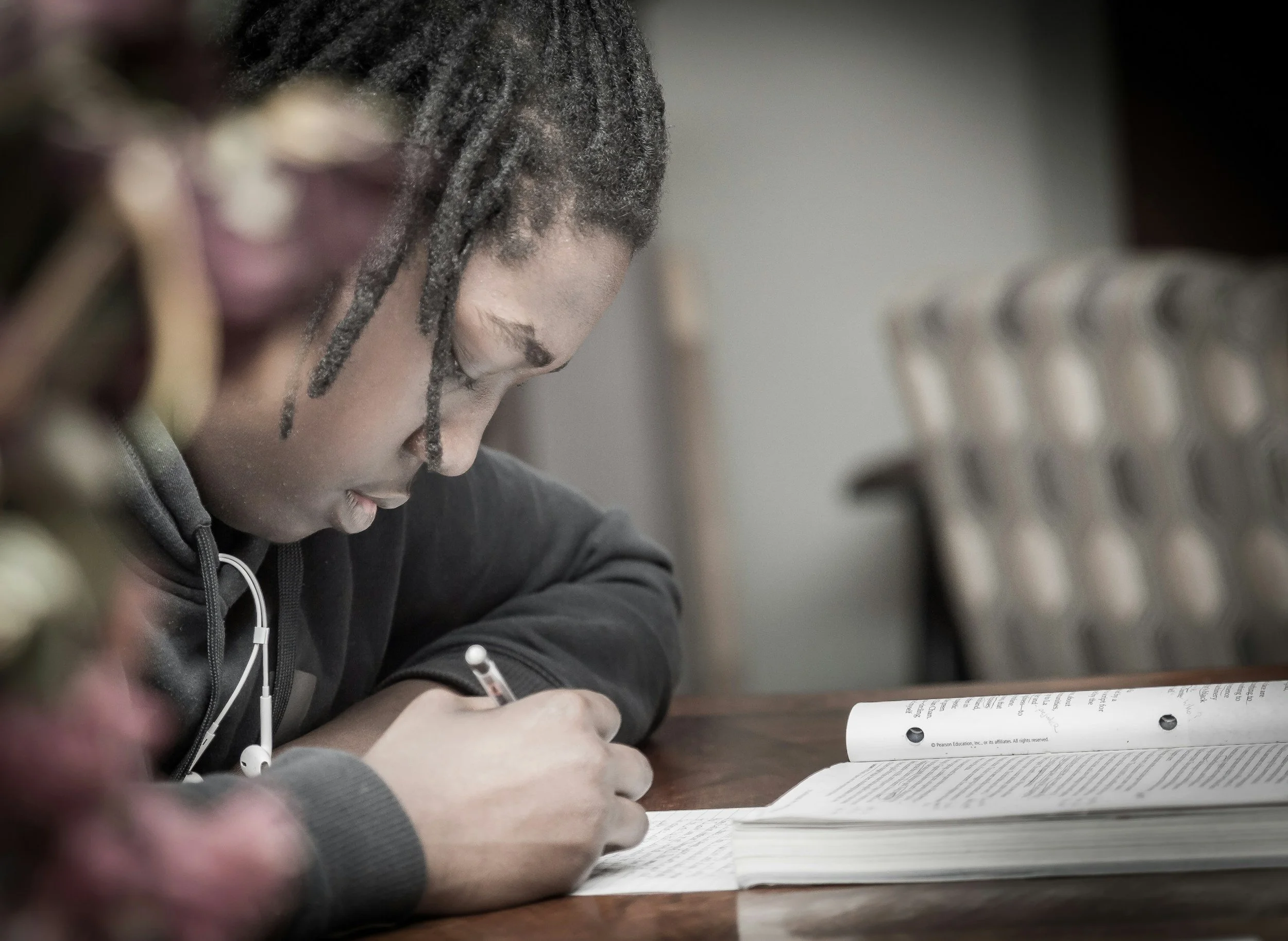 A person with dreadlocks studying with a large open book and a notebook on a wooden table.