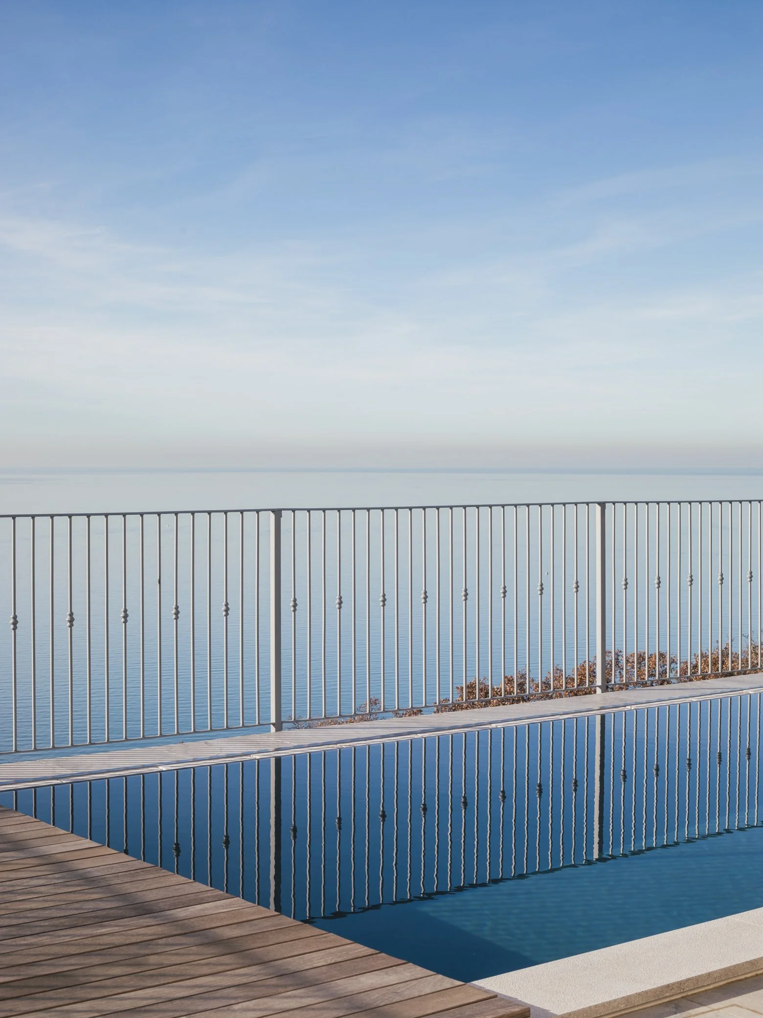 A swimming pool with reflections of a metal railing, overlooking a calm body of water under a blue sky with scattered clouds.
