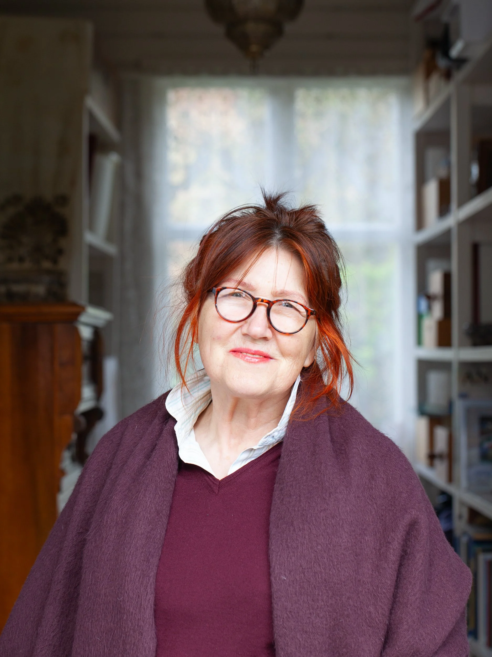 A woman with red hair, glasses, and a gentle smile, wearing a white collared shirt and a maroon sweater, sitting in a cozy room with bookshelves and a large window with sheer curtains in the background.