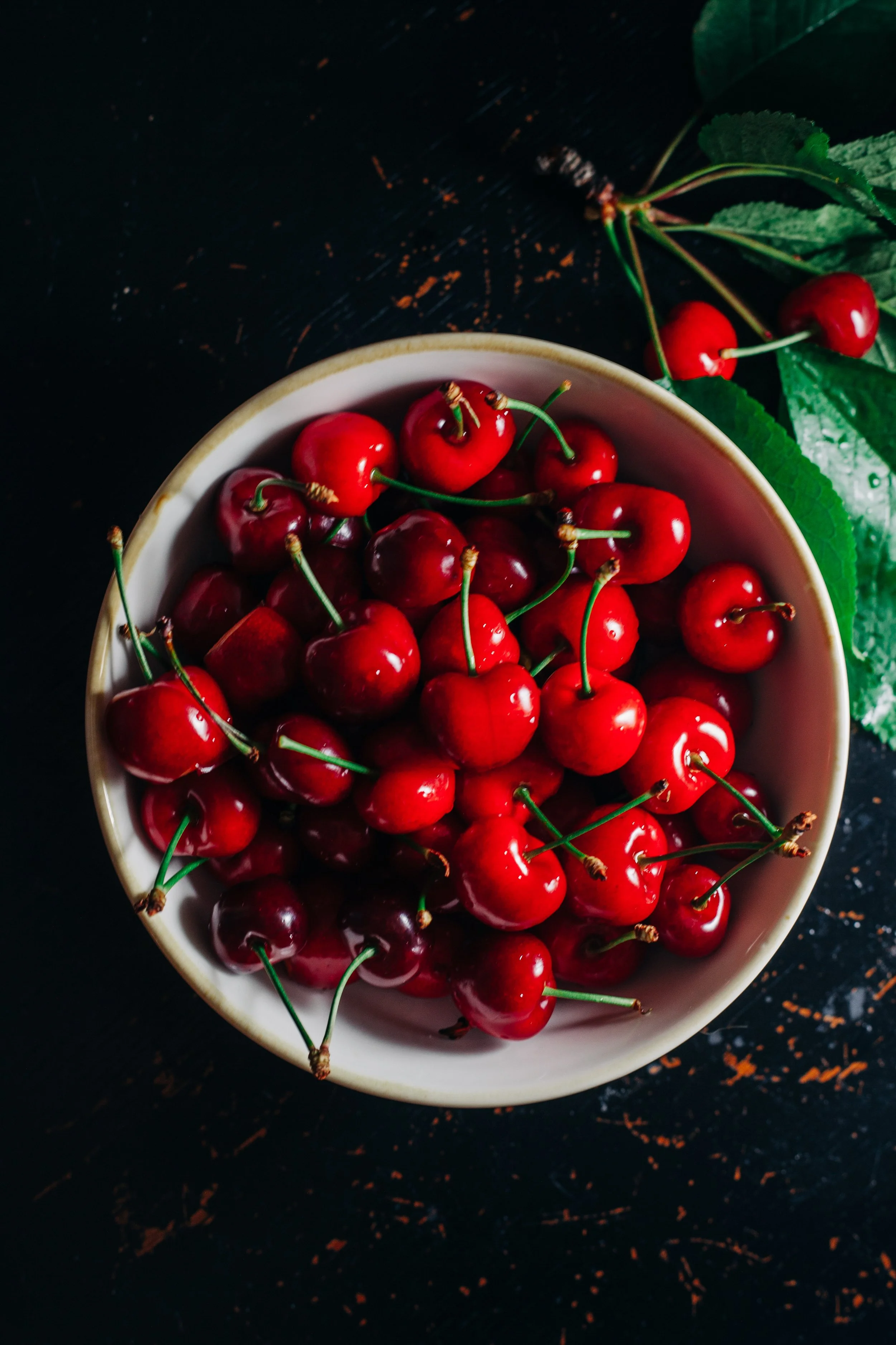 A bowl of fresh red cherries on a dark wooden surface with a few cherries and green leaves nearby.
