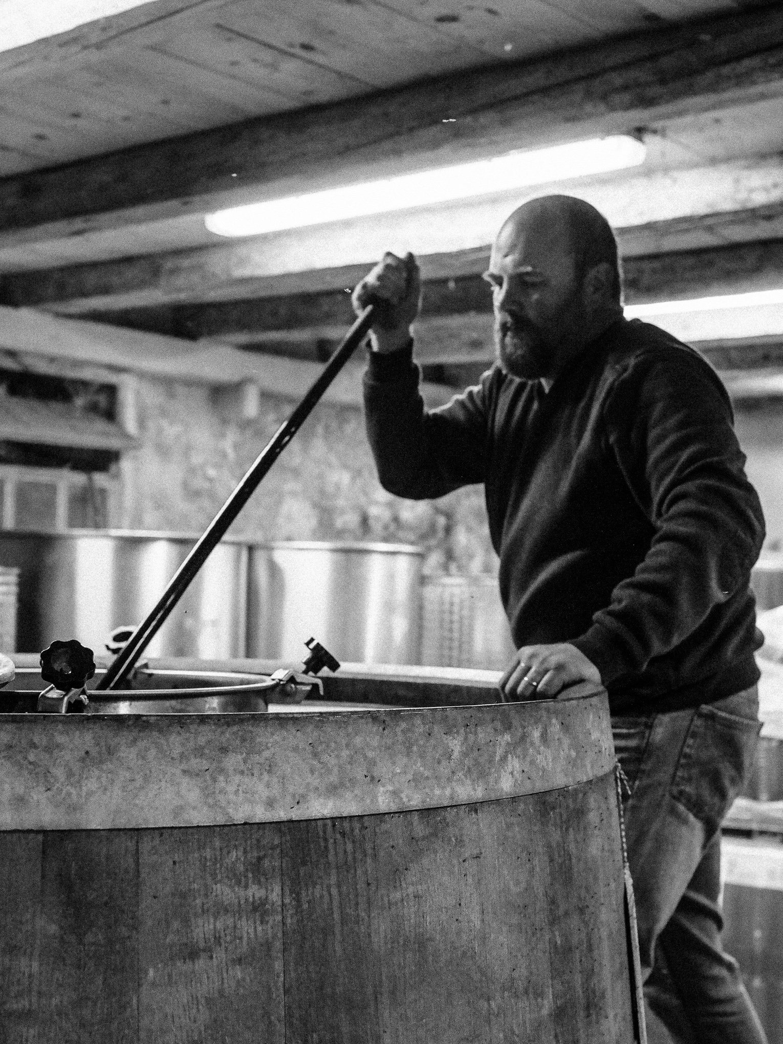 A man stirring a large vat, possibly in a brewery or winery, in an industrial setting with wooden beams and metal tanks in the background.