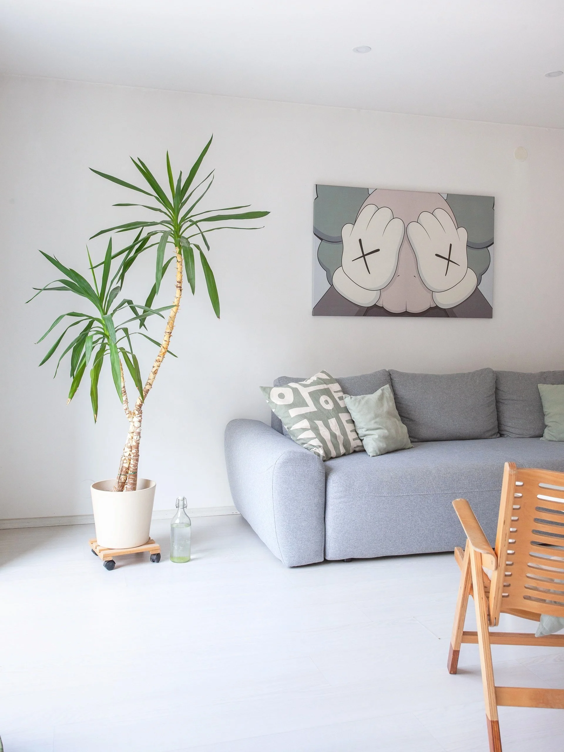 Living room with a tall potted plant on wheels, a gray sofa with patterned and plain pillows, wall art depicting a person with hands covering their eyes, a wooden chair, and a glass bottle on the floor.
