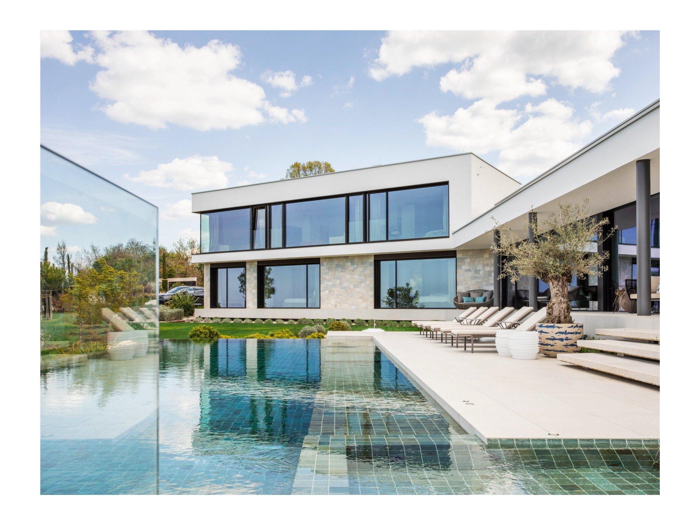 Modern house with large glass windows, outdoor pool, lounge chairs, trees, and a blue sky