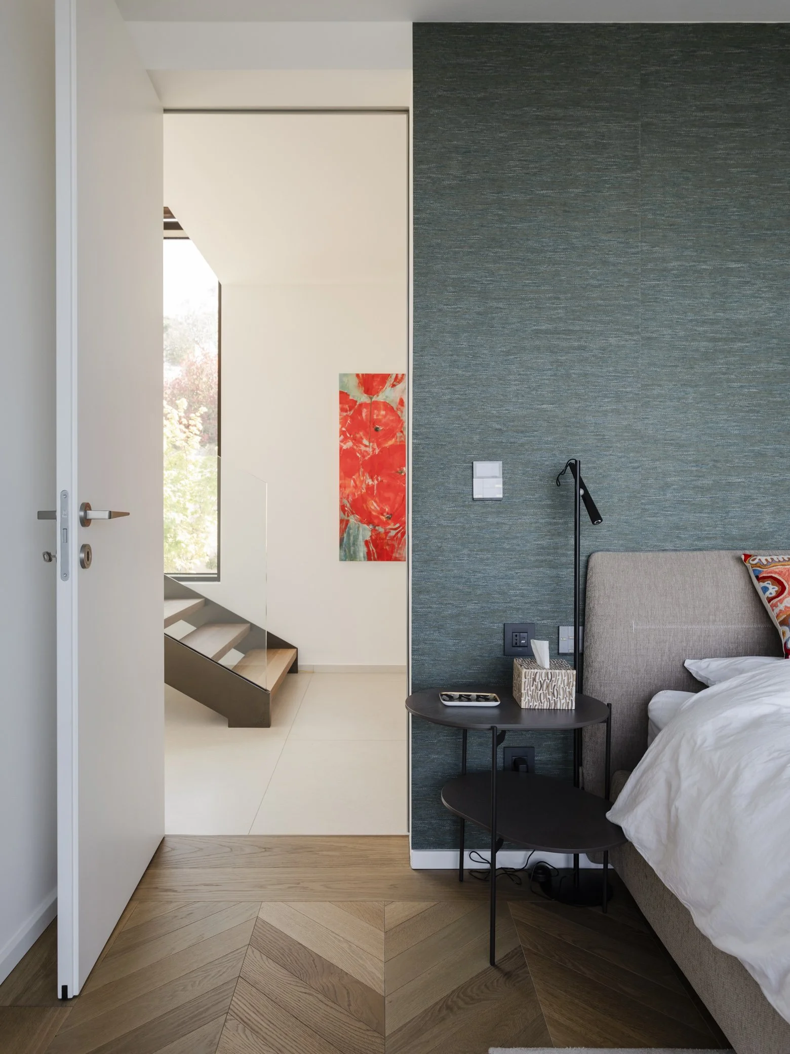 Modern bedroom with a beige bed, dark green textured accent wall, black bedside table with a tray and tissue box, wall-mounted reading lamp, and a doorway leading to a hallway with stairs and a colorful abstract painting.