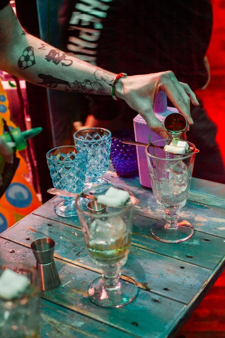 A bartender is pouring a dark liquid into a glass filled with ice and white whipped cream, with other colorful glassware and utensils on a rustic wooden table.