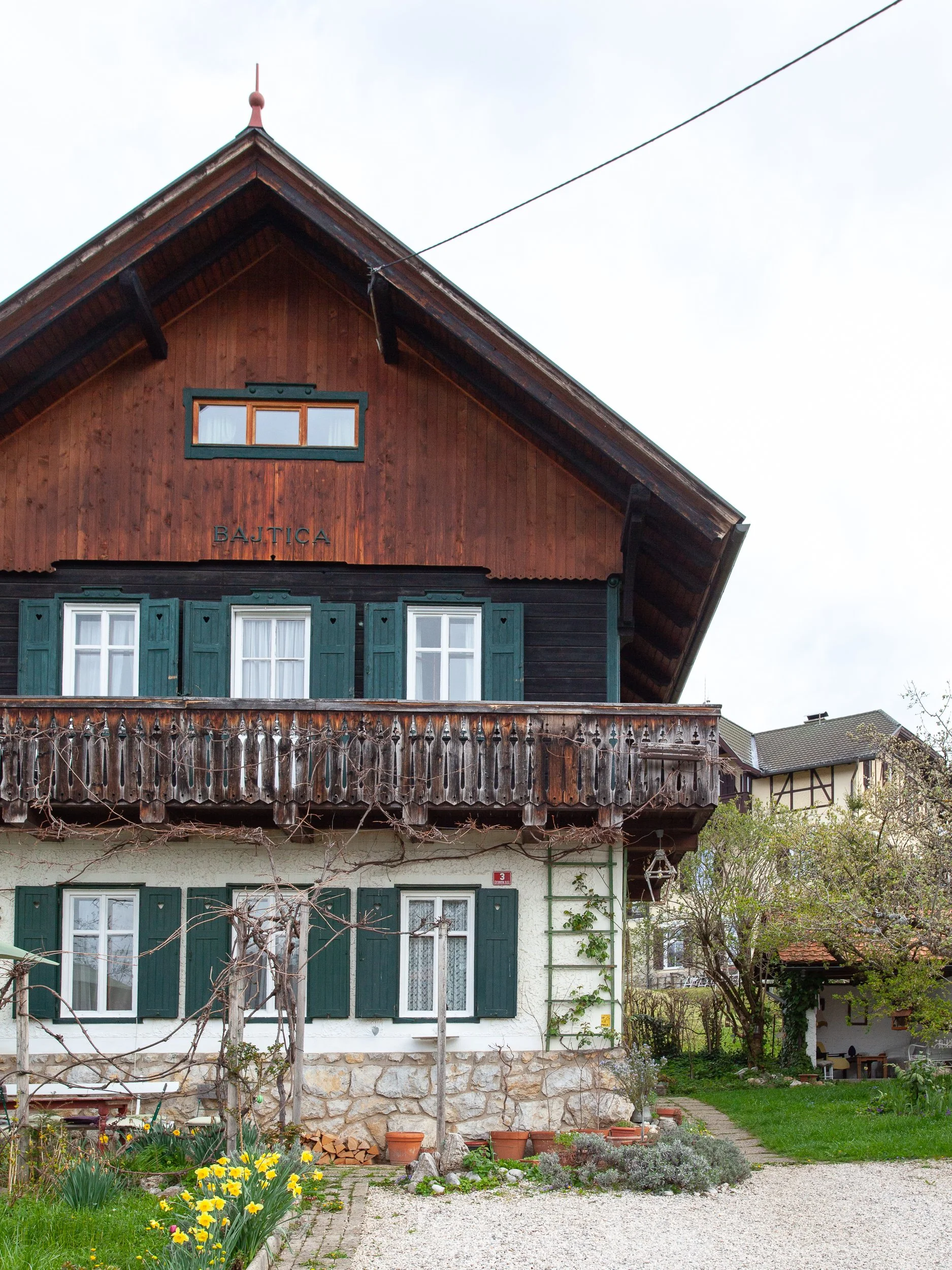 A two-story house with a wooden upper floor and a white lower floor, green shutters, a small balcony, and a garden with yellow flowers in front.