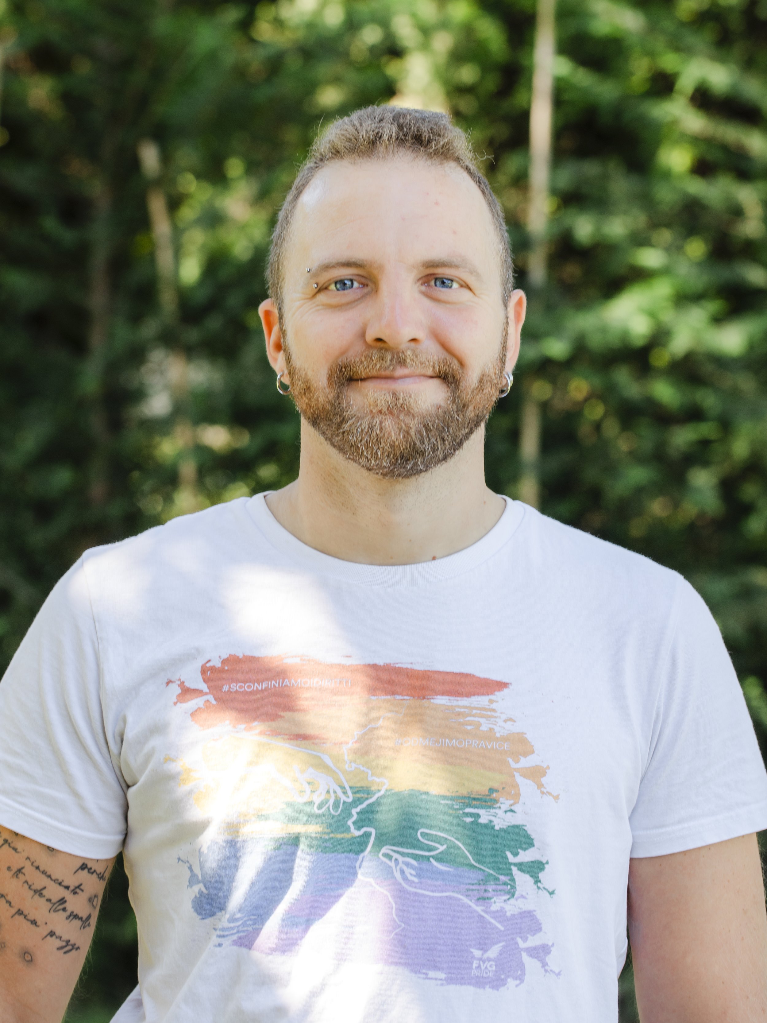 A smiling man with a beard, blue eyes, and earrings, standing outdoors in front of green trees, wearing a white t-shirt with a rainbow-colored graphic design.
