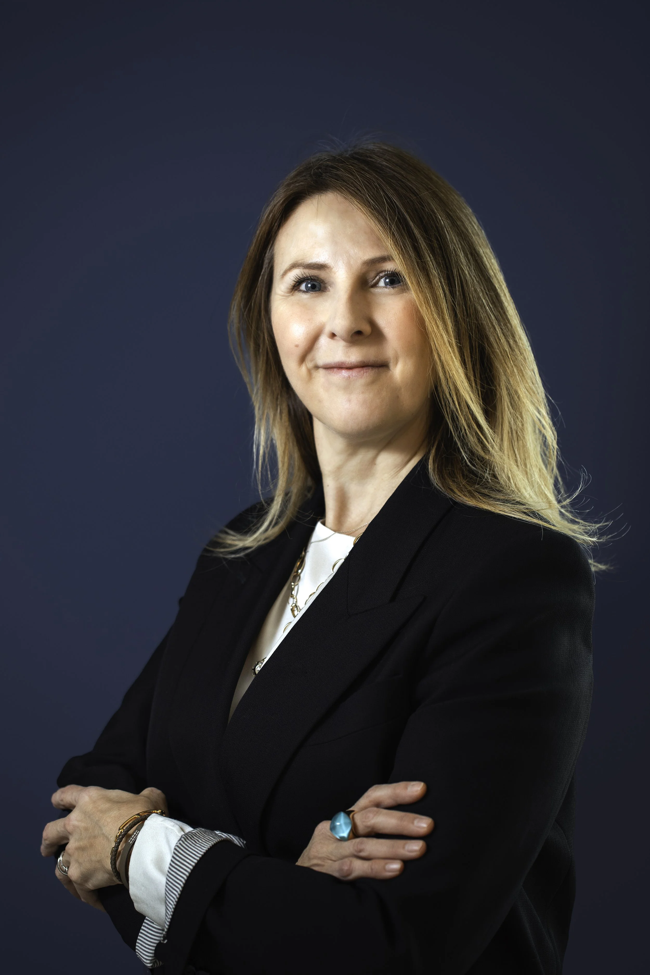 Professional woman with shoulder-length blonde hair, wearing a black blazer, white blouse, and jewelry, standing against a dark blue background, with arms crossed and a confident expression.