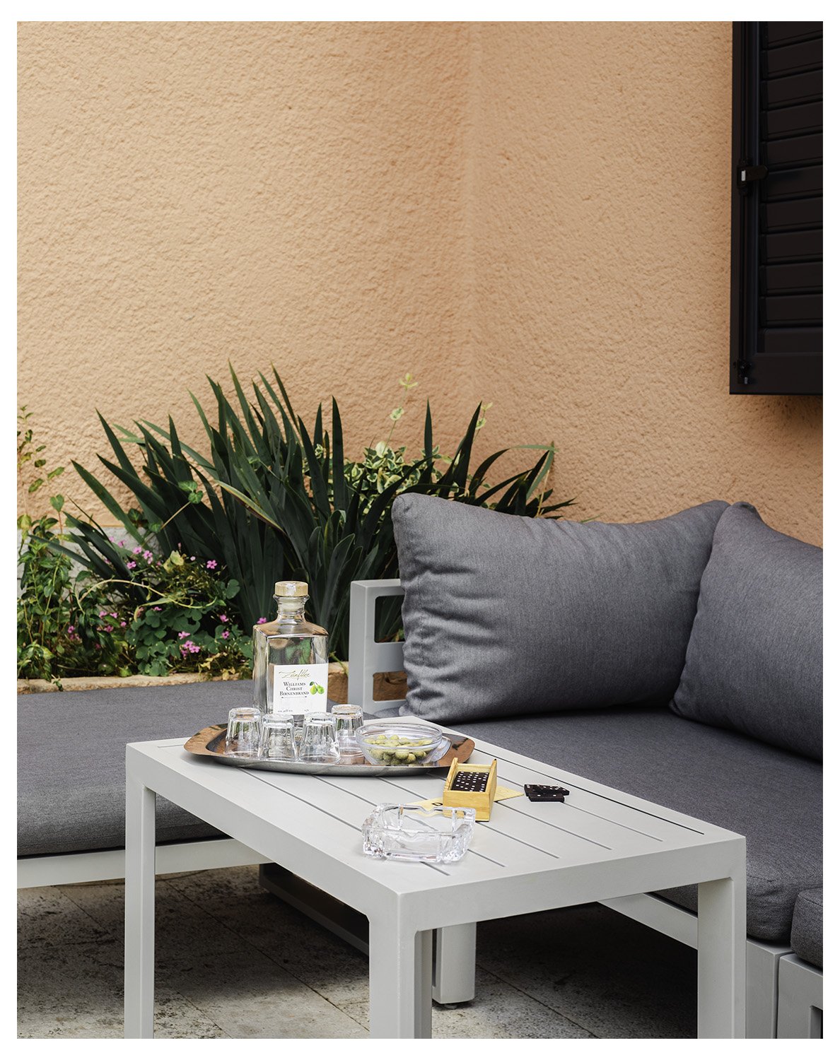 An outdoor patio scene with a white table, gray cushions, a bottle of water, glasses, a bowl of snacks, and a small game set, against a peach-colored wall with green plants and a black window shutter.