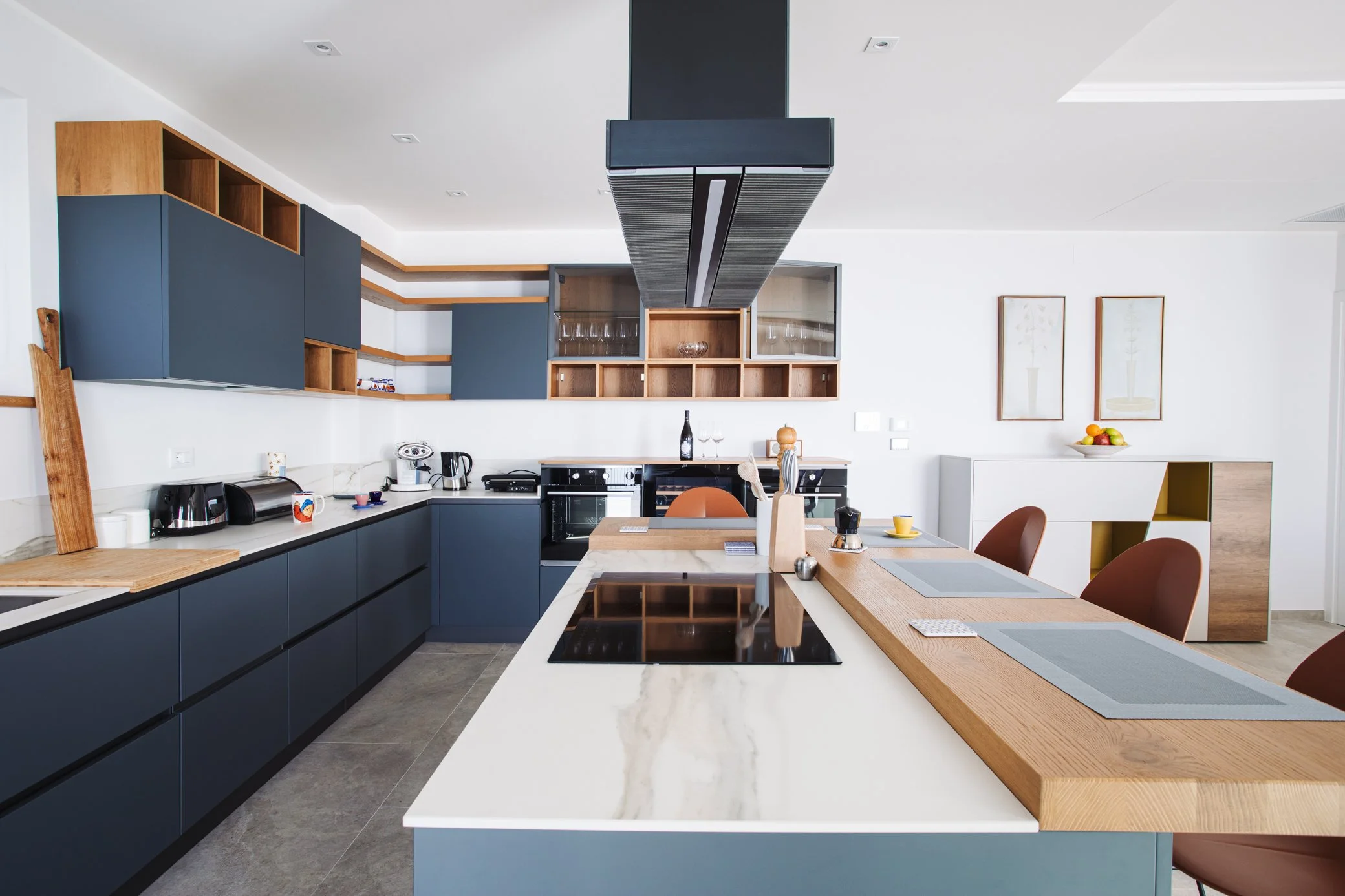 Modern kitchen with navy blue cabinets, white marble countertops, and a wooden kitchen island with placemats. Includes built-in appliances, open shelving with glasses, and a white cabinet with a bowl of fruit in the background.