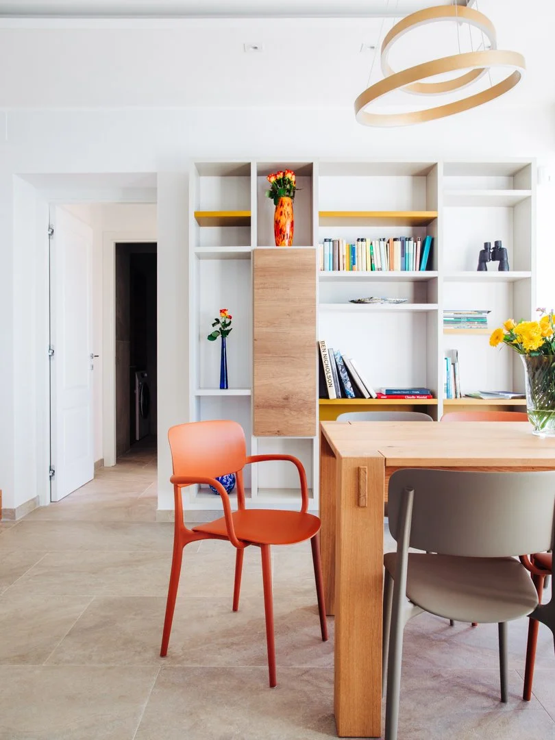 Modern dining room with a wooden table, colorful chairs, and a white bookshelf filled with books and decorative vases with flowers.