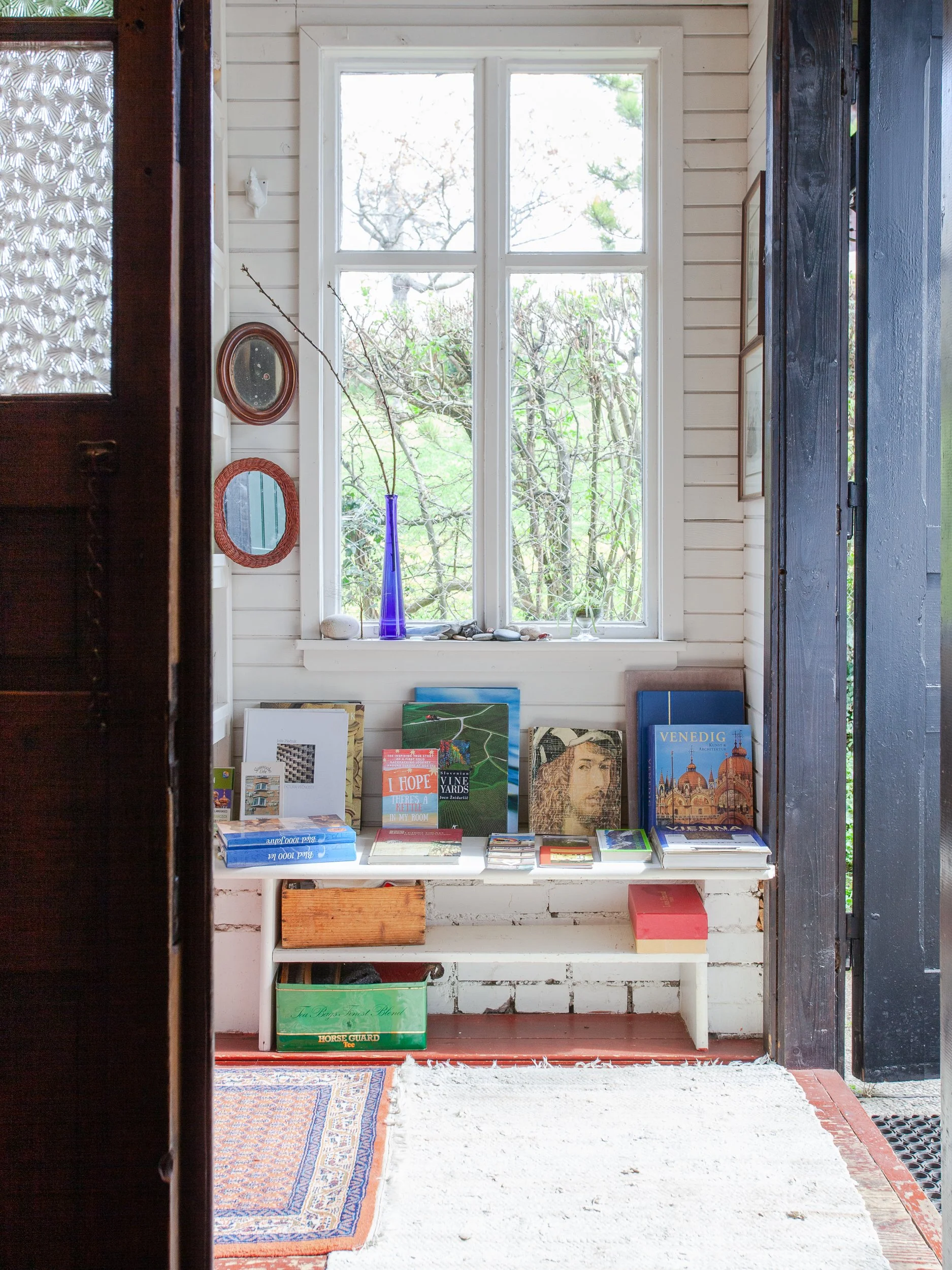 Interior of a house with a white wooden bench holding books and magazines, a tall blue glass vase on the windowsill, and small mirrors on the wall to the left of a window. The room has an open door with black trim, and a white rug on the floor.