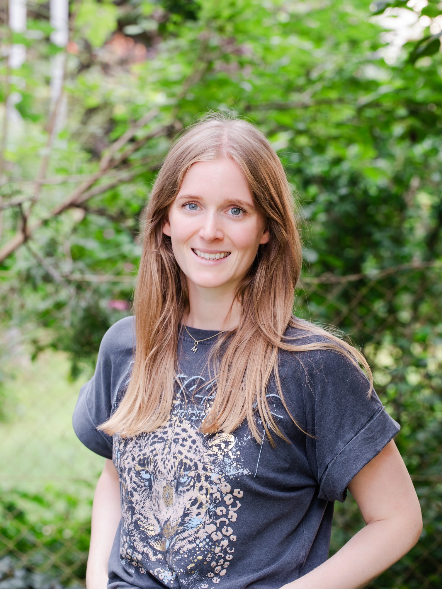 A young woman with long, light brown hair and blue eyes smiling outdoors in a green, leafy setting.