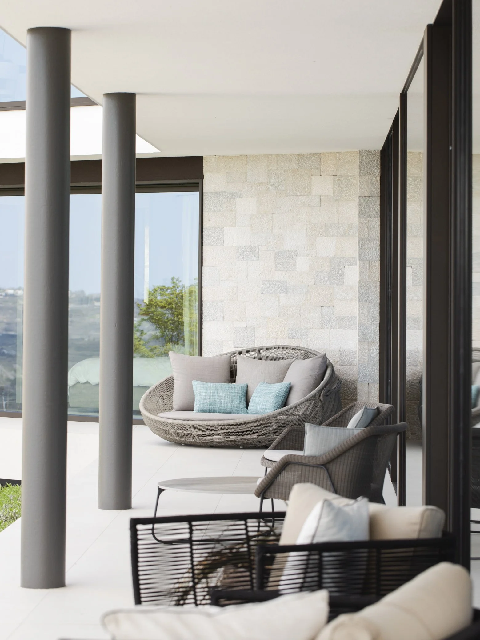 Modern patio with beige cushioned outdoor furniture, including a woven round loveseat with pillows, a wicker chair with pillows, and a white sofa with pillows, next to large glass sliding doors and windows, with a stone wall and trees outside.