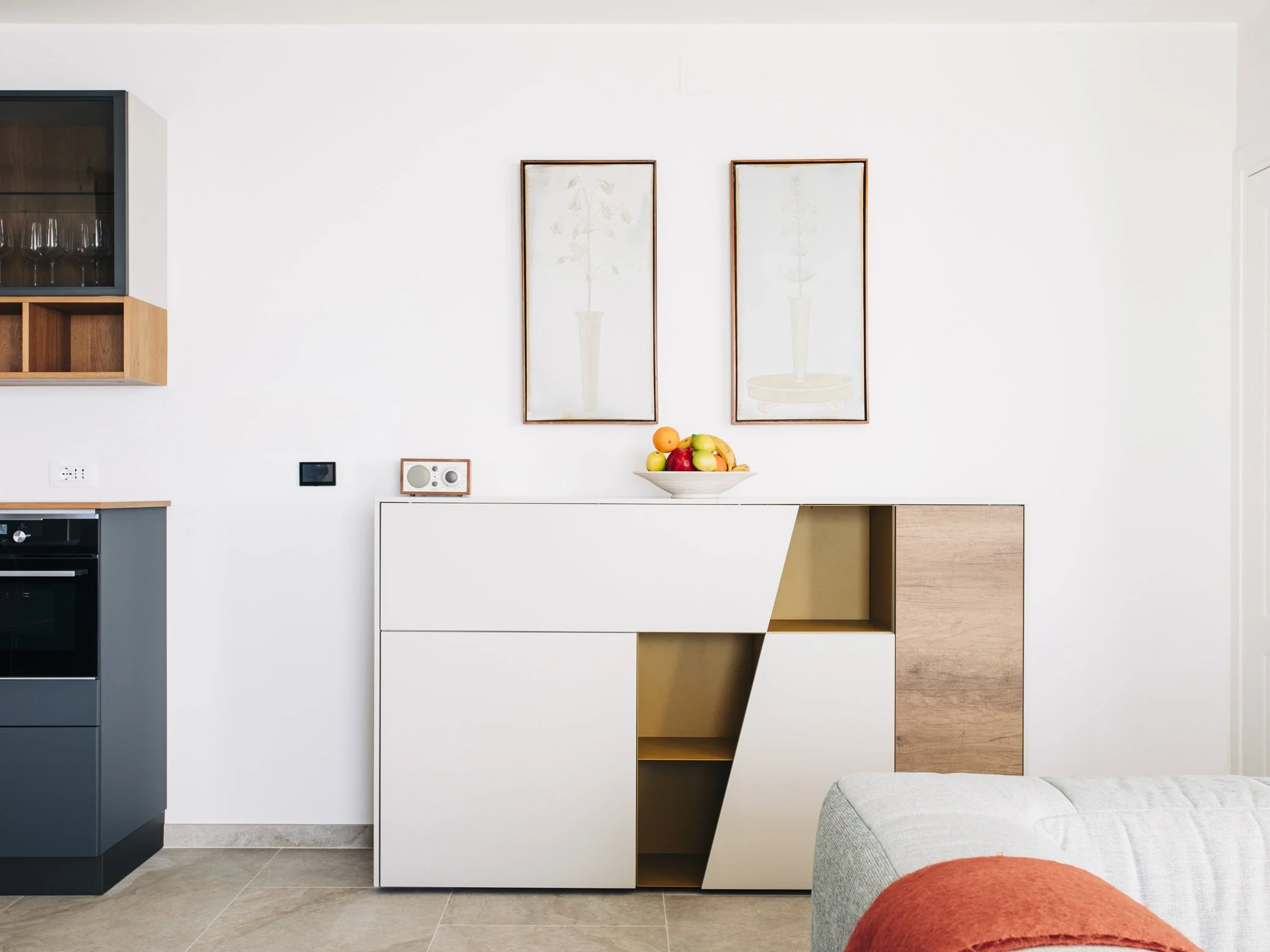 Modern interior with white and wooden furniture, framed art, and a bowl of fruit on a white and wood sideboard. Part of a gray sofa with an orange throw is visible in the foreground.
