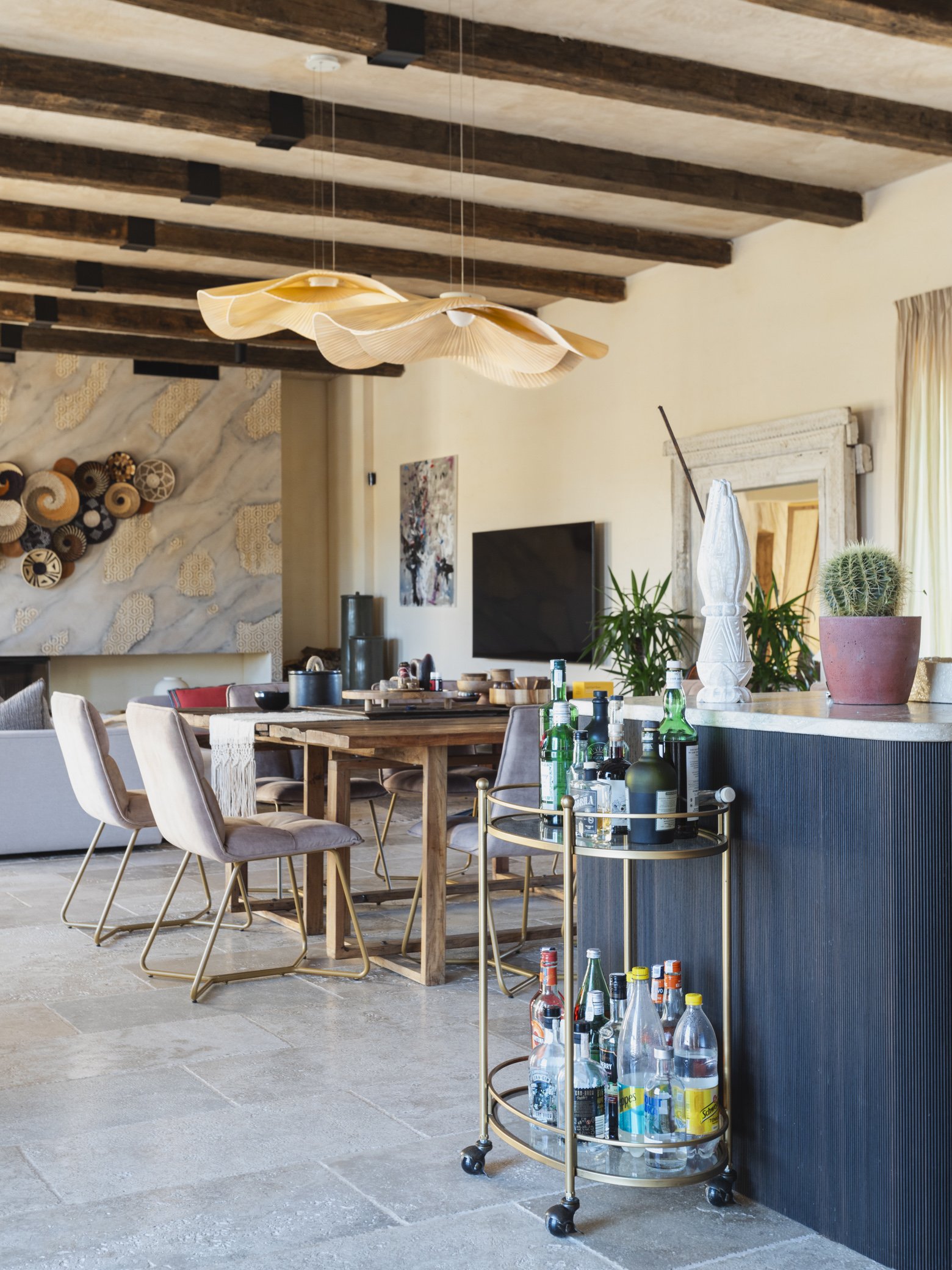 Modern dining room with wooden ceiling beams, a wooden dining table, pink upholstered chairs, a bar cart with liquor bottles, and abstract wall art.