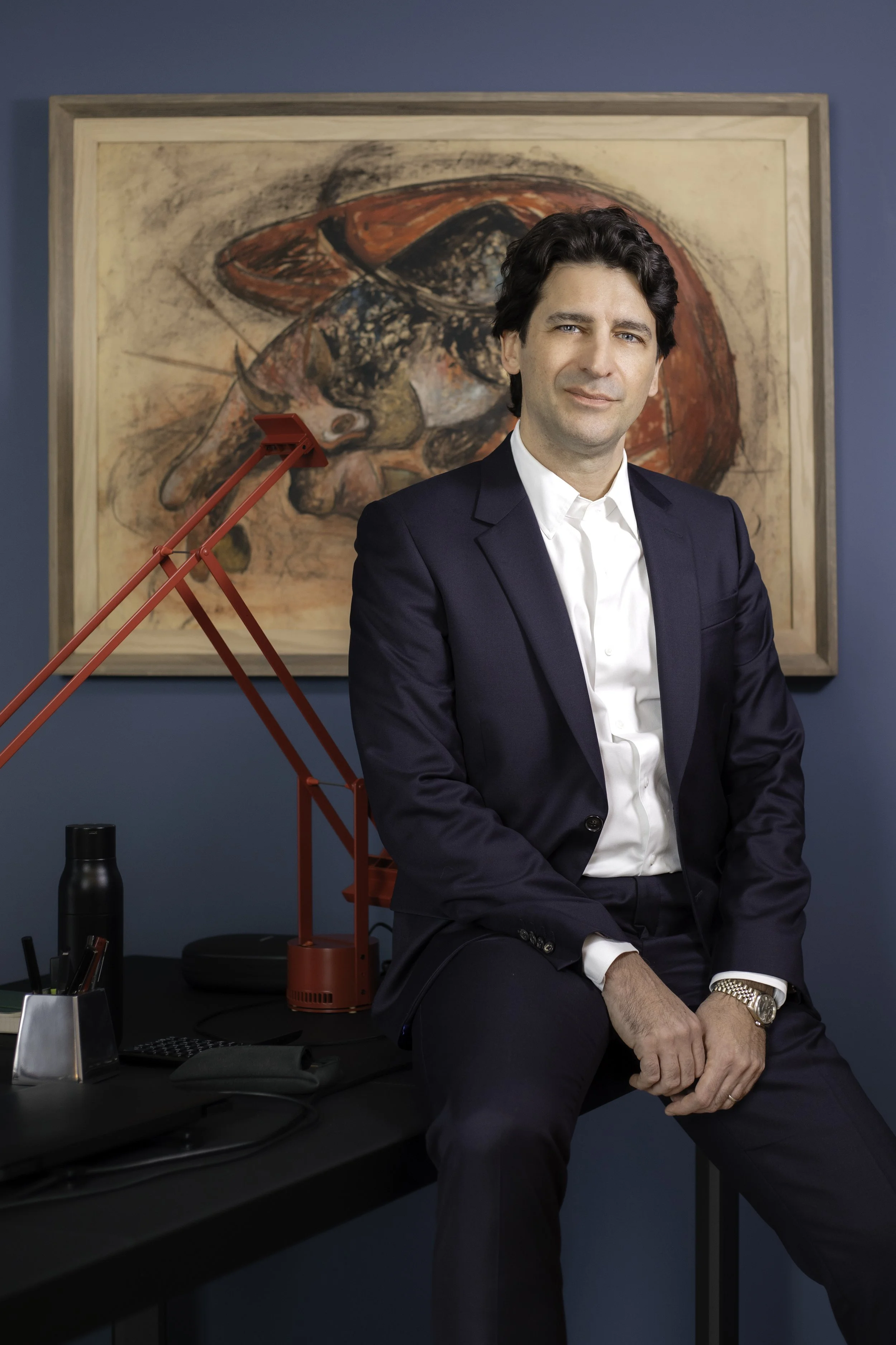 A man in a dark navy suit and white shirt sitting on the edge of a black desk in an office, with a red desk lamp and a framed abstract painting in the background.