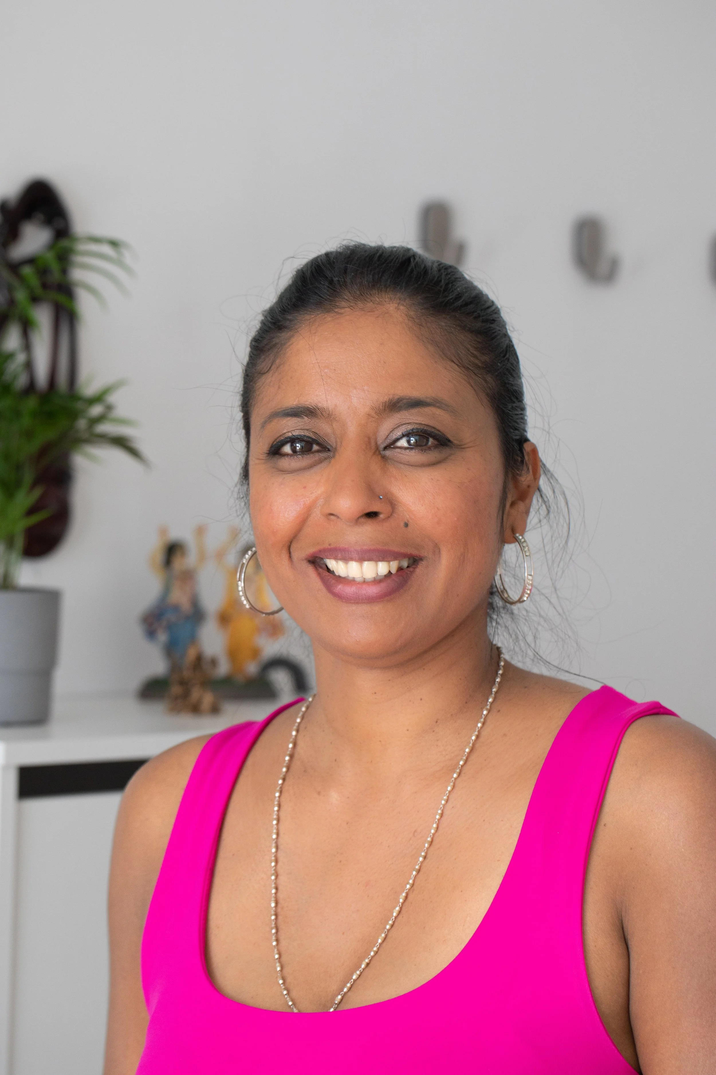 Smiling woman in a pink top with jewelry, standing indoors with a potted plant and Indian figurines in the background.