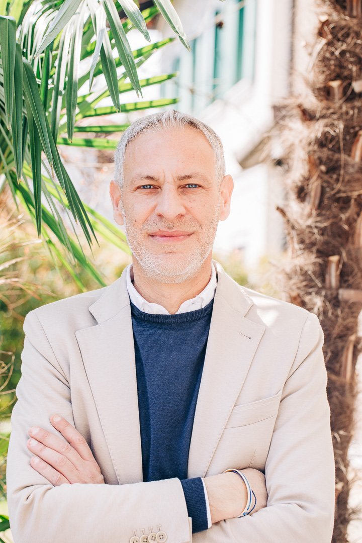 A middle-aged man with gray hair and a beard standing outdoors with arms crossed, wearing a beige blazer over a dark sweater and white collared shirt, with green foliage and palm trees in the background.