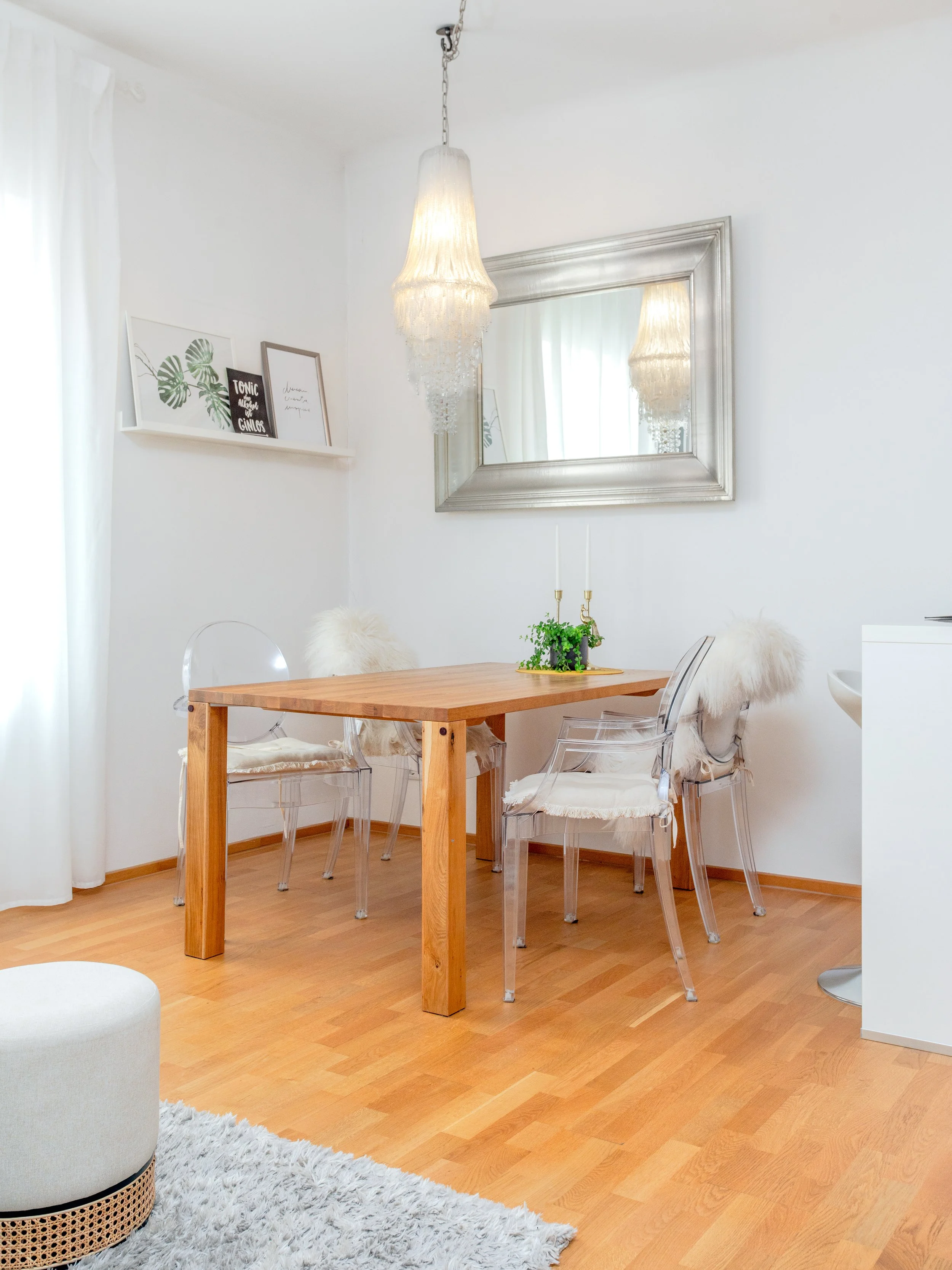 A dining room with a wooden table, transparent chairs with white furry cushions, a decorative mirror, framed art prints, and a hanging chandelier.