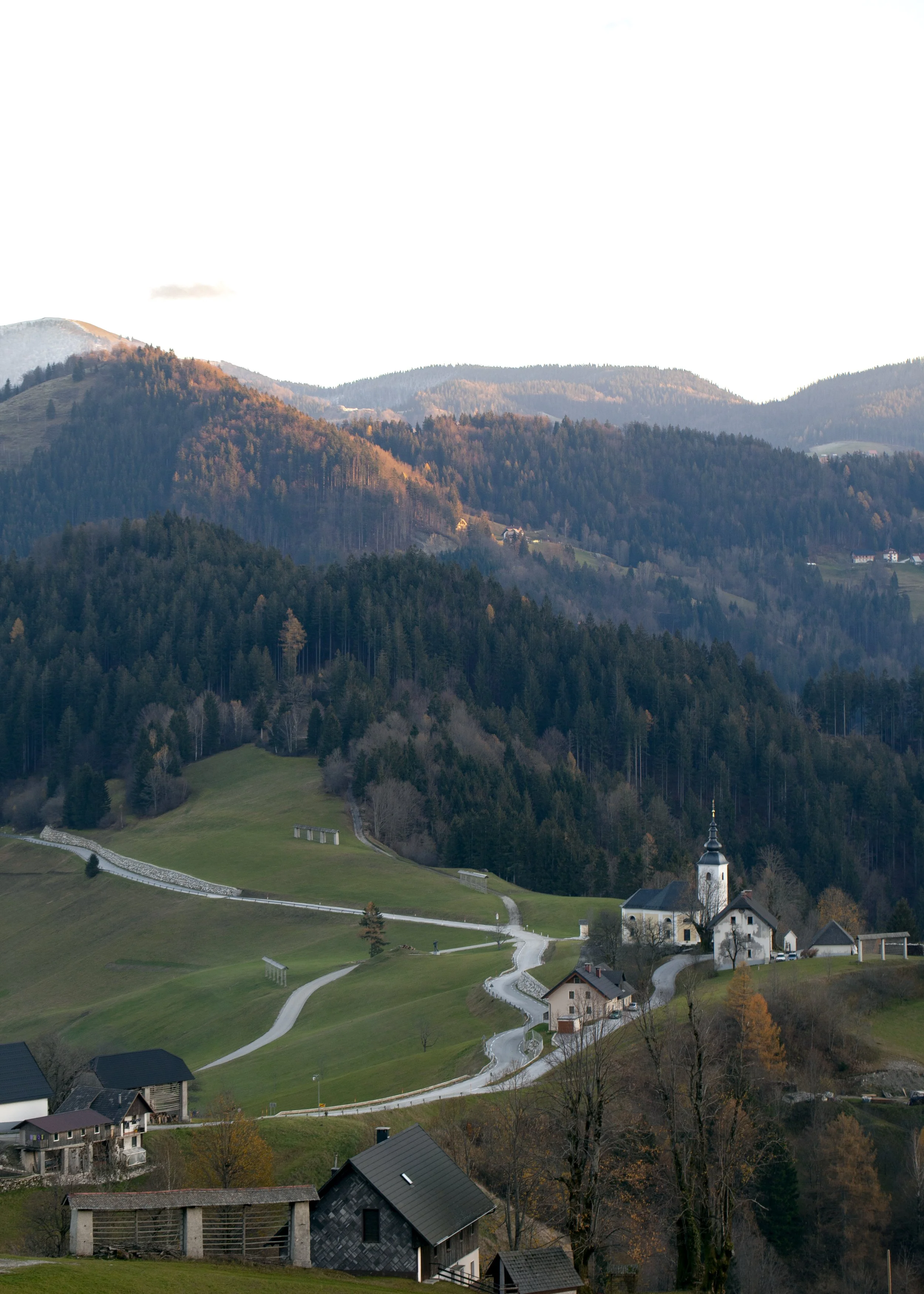 Scenic view of a hilly landscape with a winding road leading to a small church with a steeple, surrounded by houses, trees, and forested mountains in the background during late afternoon or early evening.