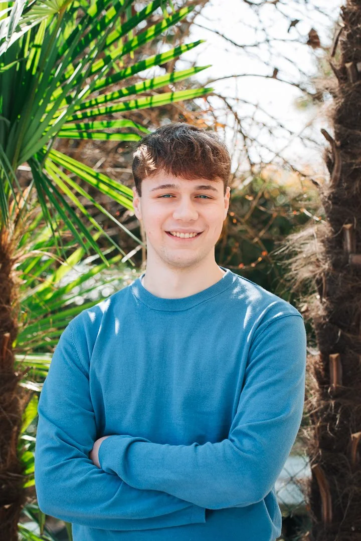 A young man with brown hair, wearing a blue long-sleeve shirt, smiling with arms crossed, standing outdoors in front of green palm leaves and a tree trunk.