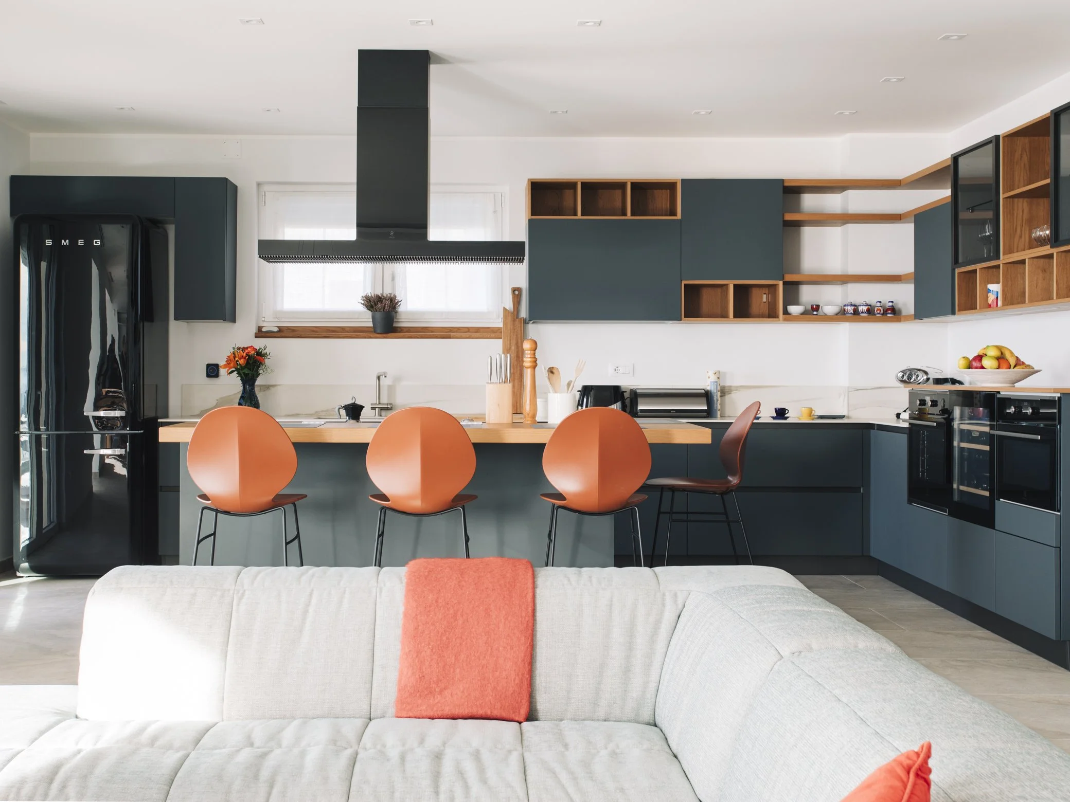 Modern kitchen with teal cabinets, orange chairs at a wooden island, black refrigerator, and a beige sofa with orange pillow in foreground.