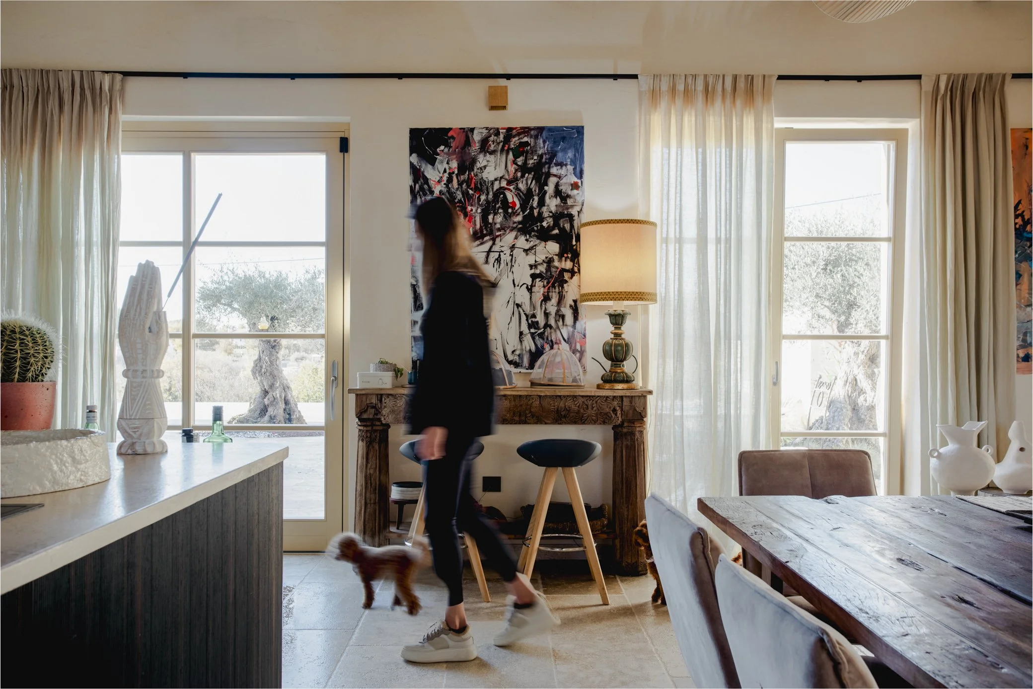 A woman walking inside a living room with two small dogs, with large windows, sheer curtains, artwork, a wooden table, and decorative items.