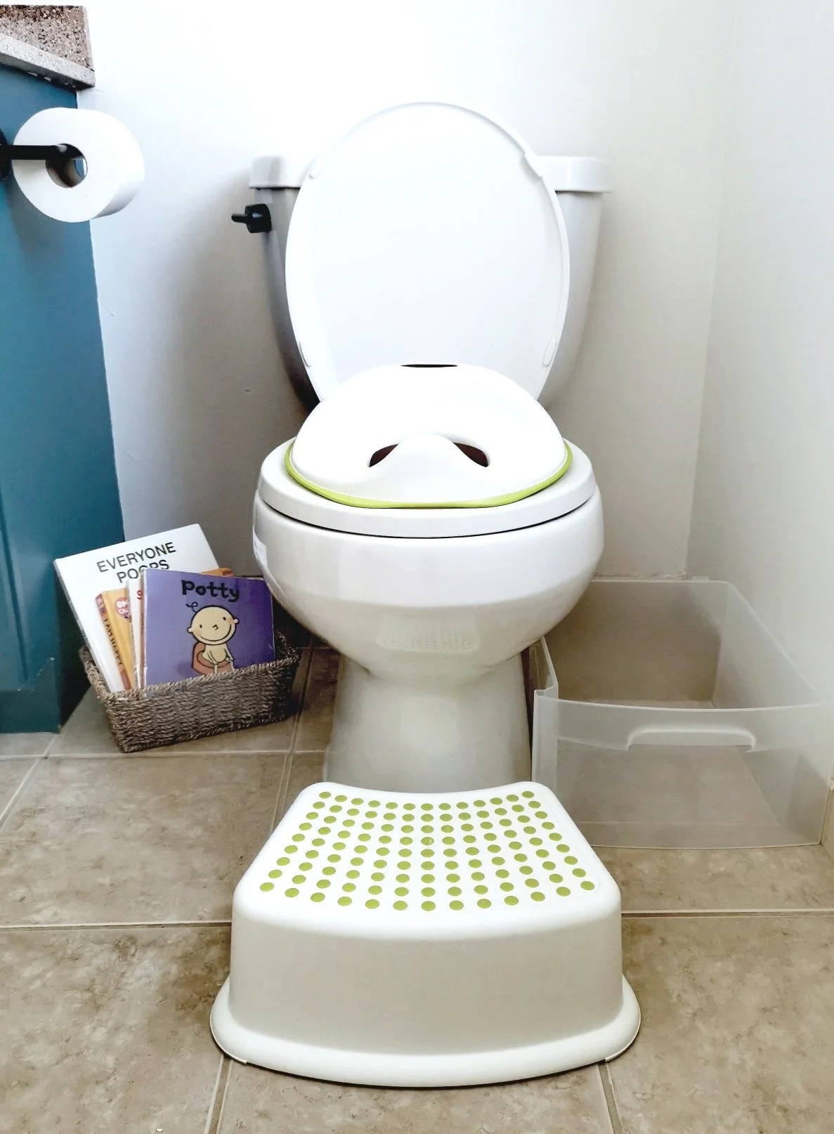 A toilet with the lid open. A toilet adapter sits on the toilet seat. Left of the toilet is a basket of children's books about toileting. In front of the toilet is a footstool. Right of the toilet is an empty plastic container.