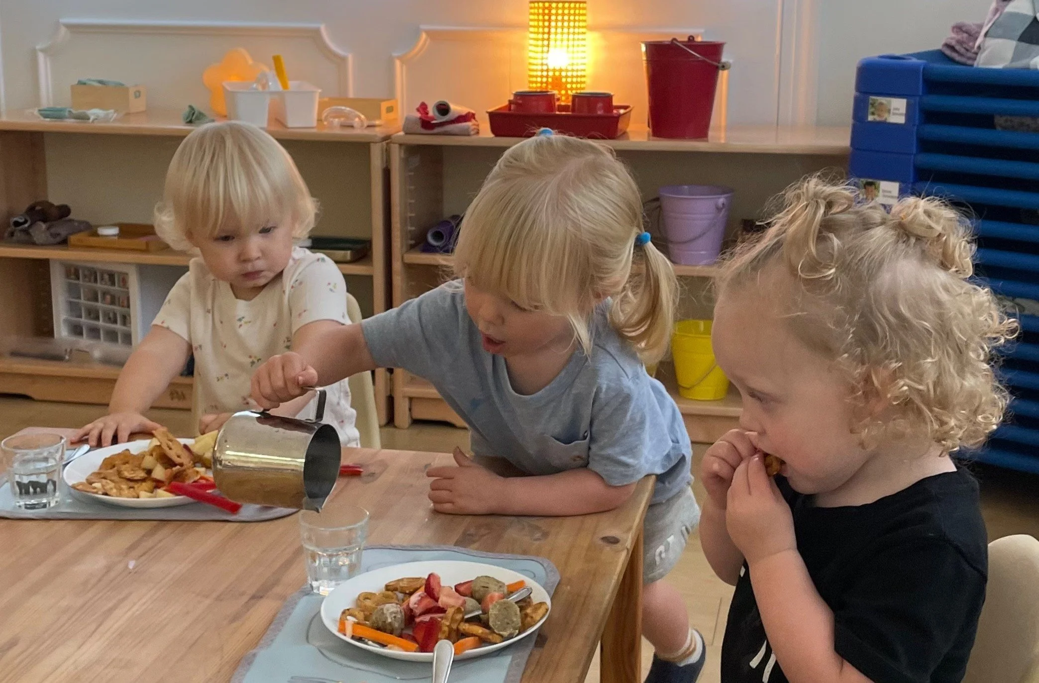 A two year old child is carefully pouring water for two toddlers who are seated at a table. The child serving is using a pitcher to pour the water into the toddler's cup.