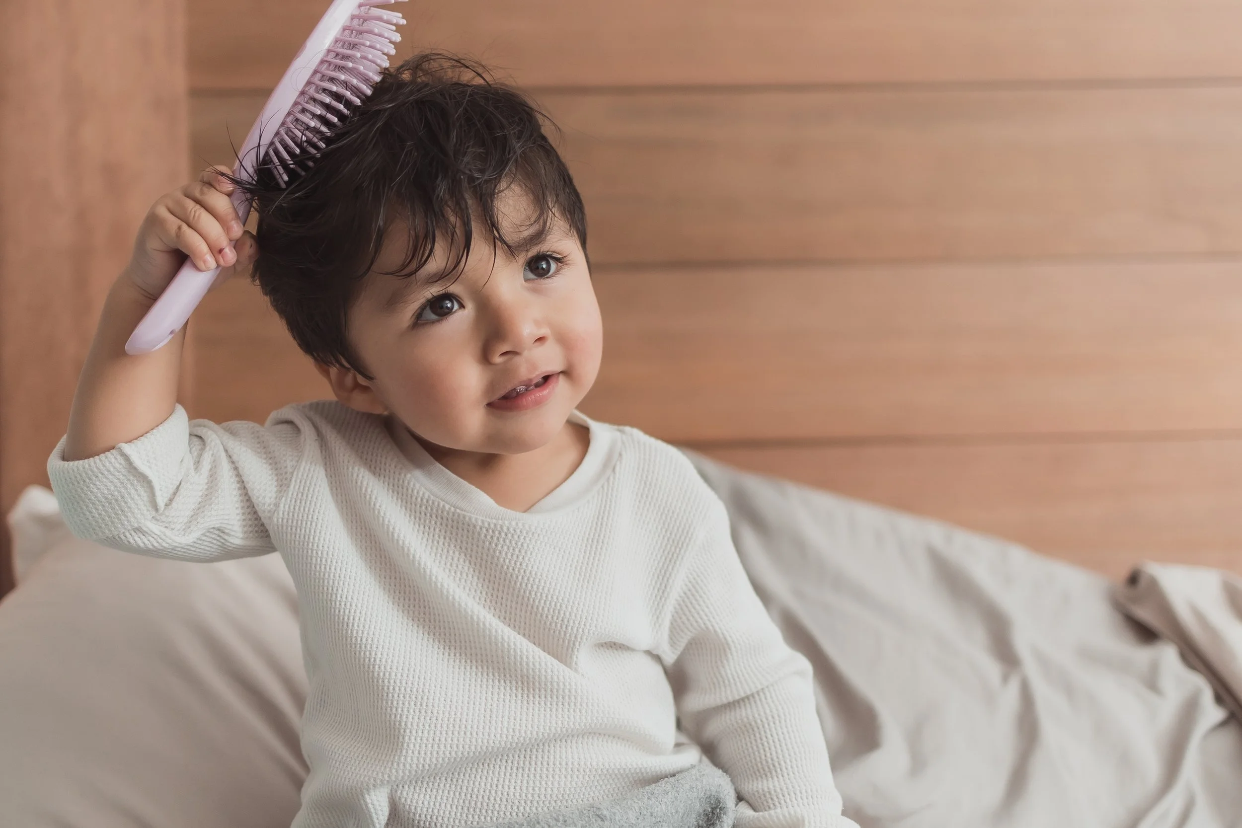A toddler wearing loose white pajamas, holding an adult-sized pink hairbrush against his or her hair, seated among white pillows in front of two wooden walls.