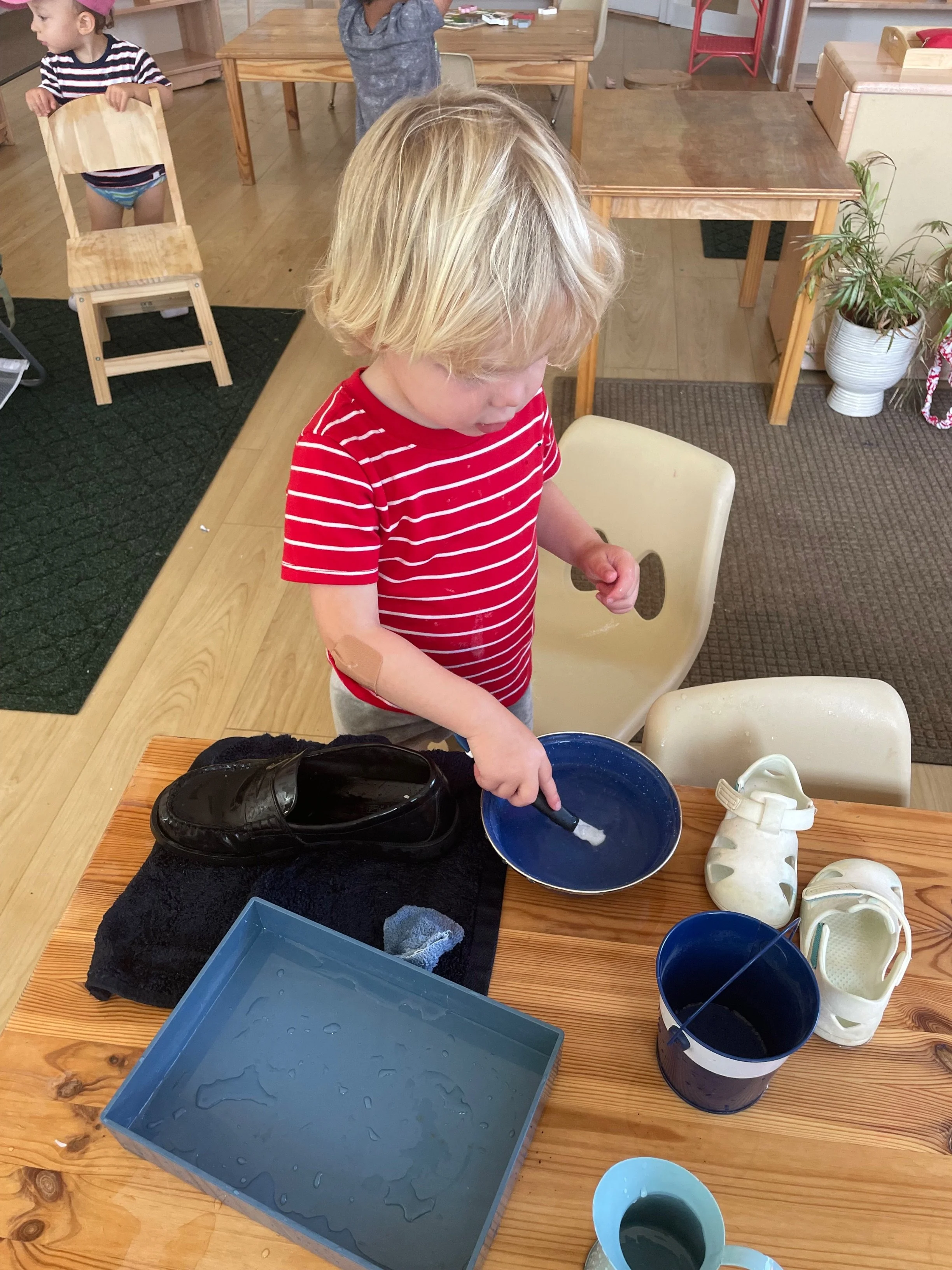 When a child chooses shoe washing work they use a pitcher to bring water from the sink. They pour the water in a bowl, wet the brush, wash their shoes, pour the bowl of water into a bucket, and empty the bucket into the sink.