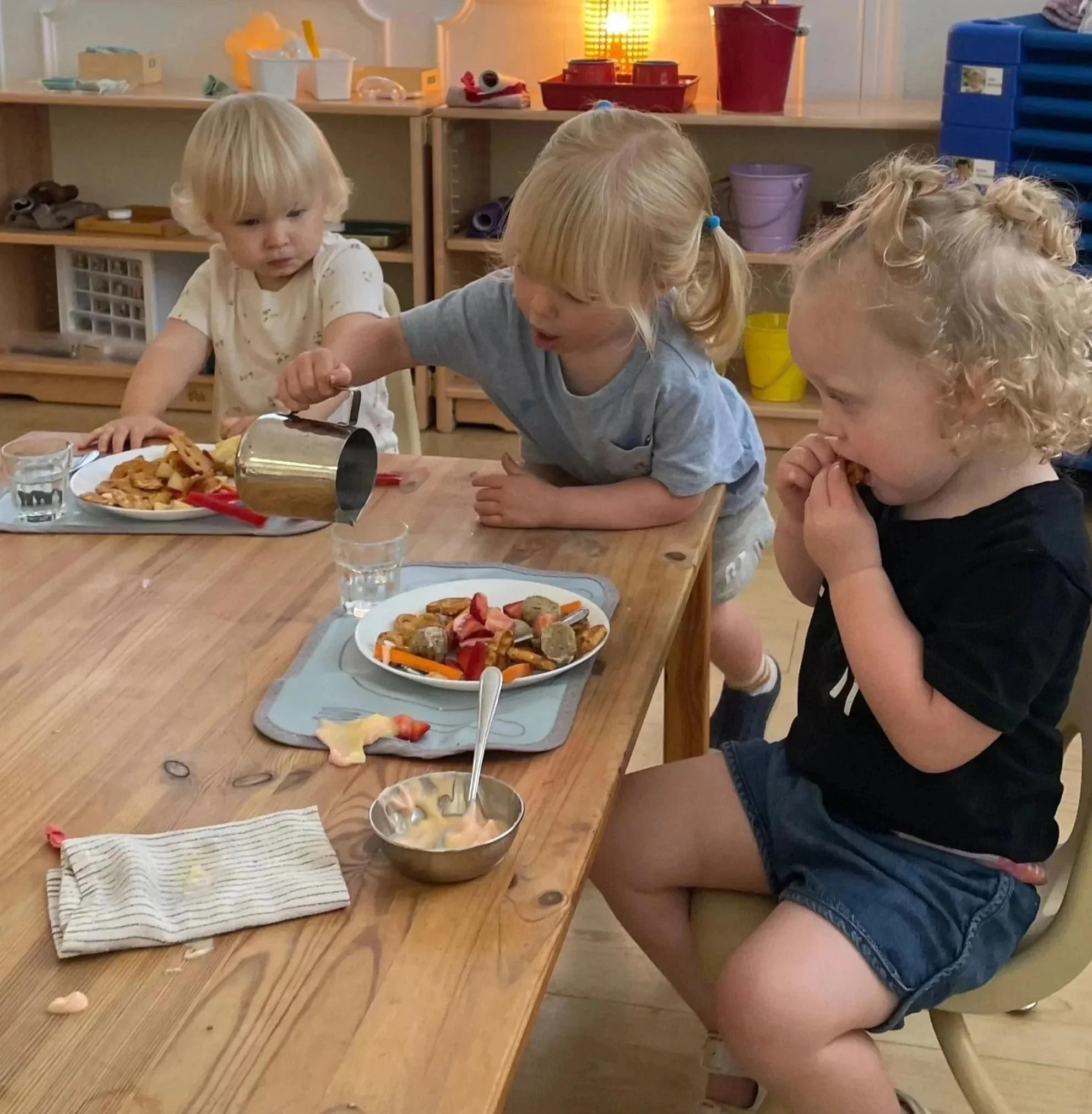 A two year old child is carefully pouring water for two toddlers who are seated at a table. The child serving is using a pitcher to pour the water into the toddler's cup.