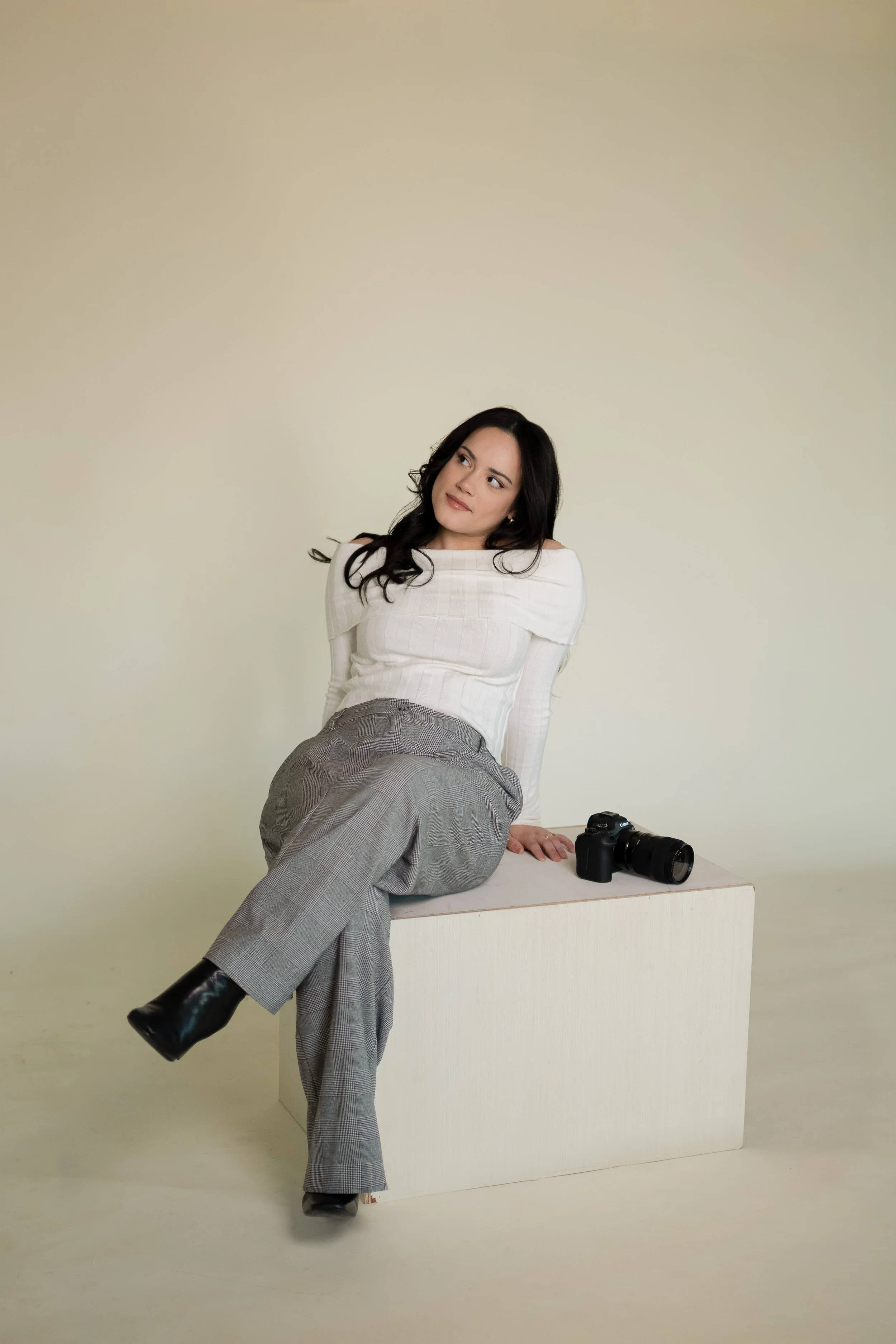 A woman with long dark hair sitting on a white block next to a camera against a plain light-colored background.