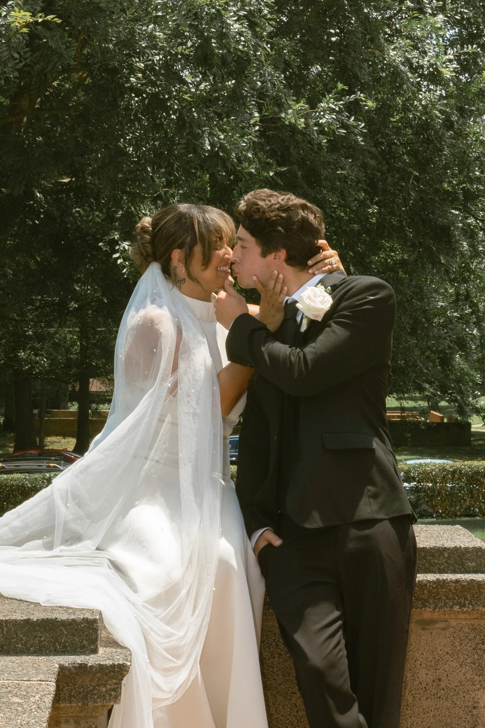 A bride and groom sharing an intimate moment outdoors, with the groom gently holding the bride's face as they lean in for a kiss, surrounded by green trees.