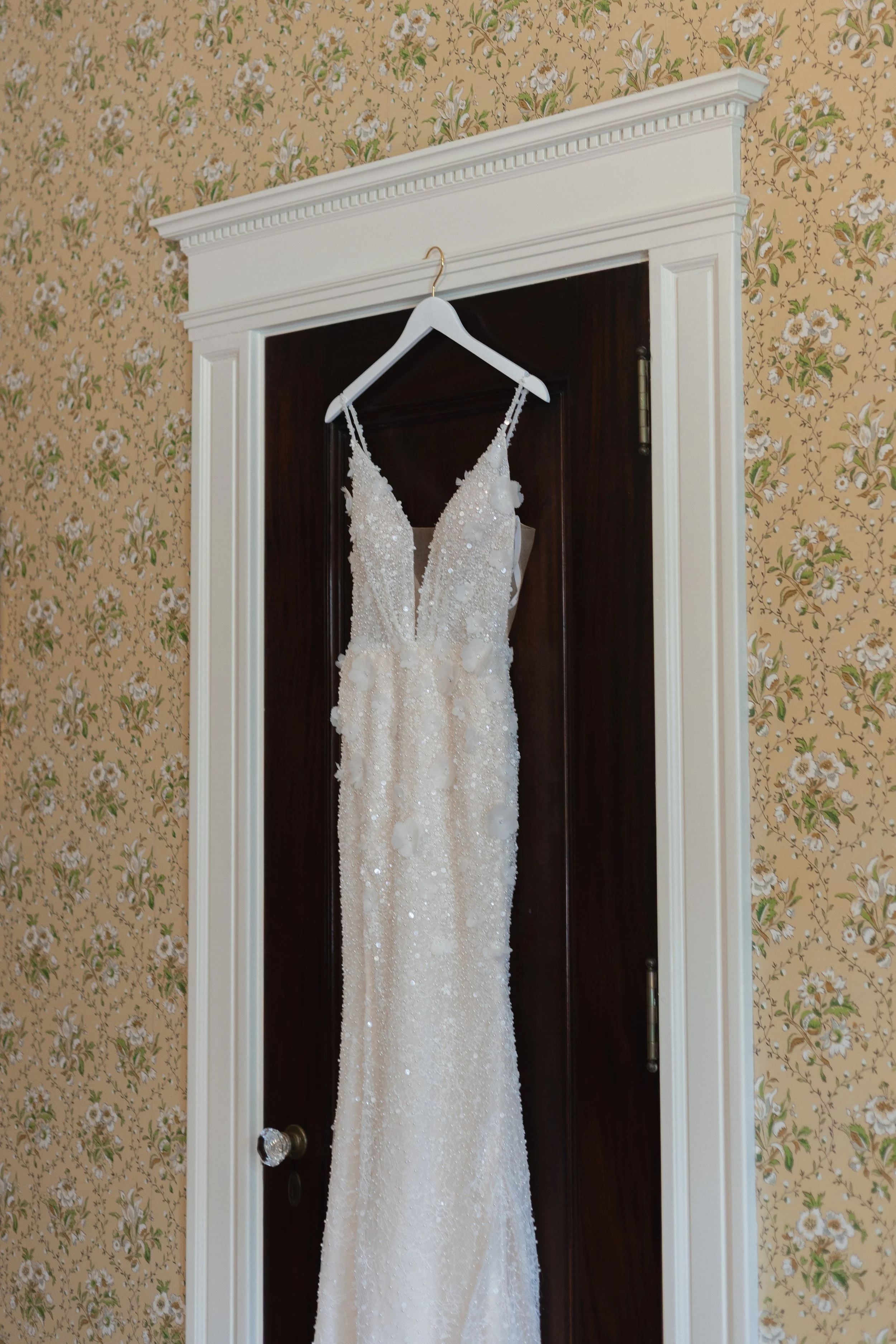 Wedding dress with intricate beadwork and floral applique hanging on a white hanger in front of a dark wooden door, with floral wallpaper in the background.