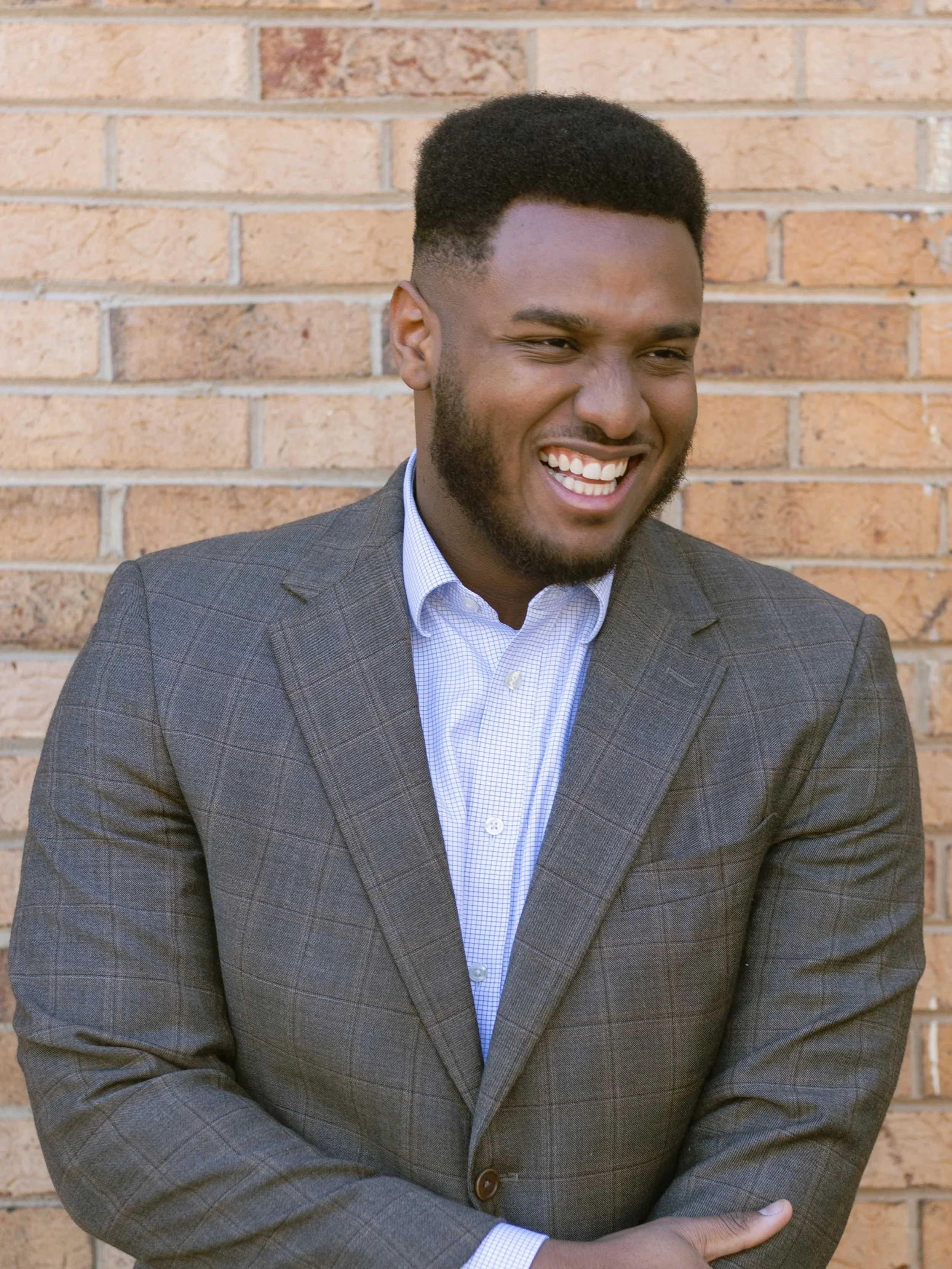 A smiling Black man in a gray plaid blazer and white dress shirt standing against a brick wall.
