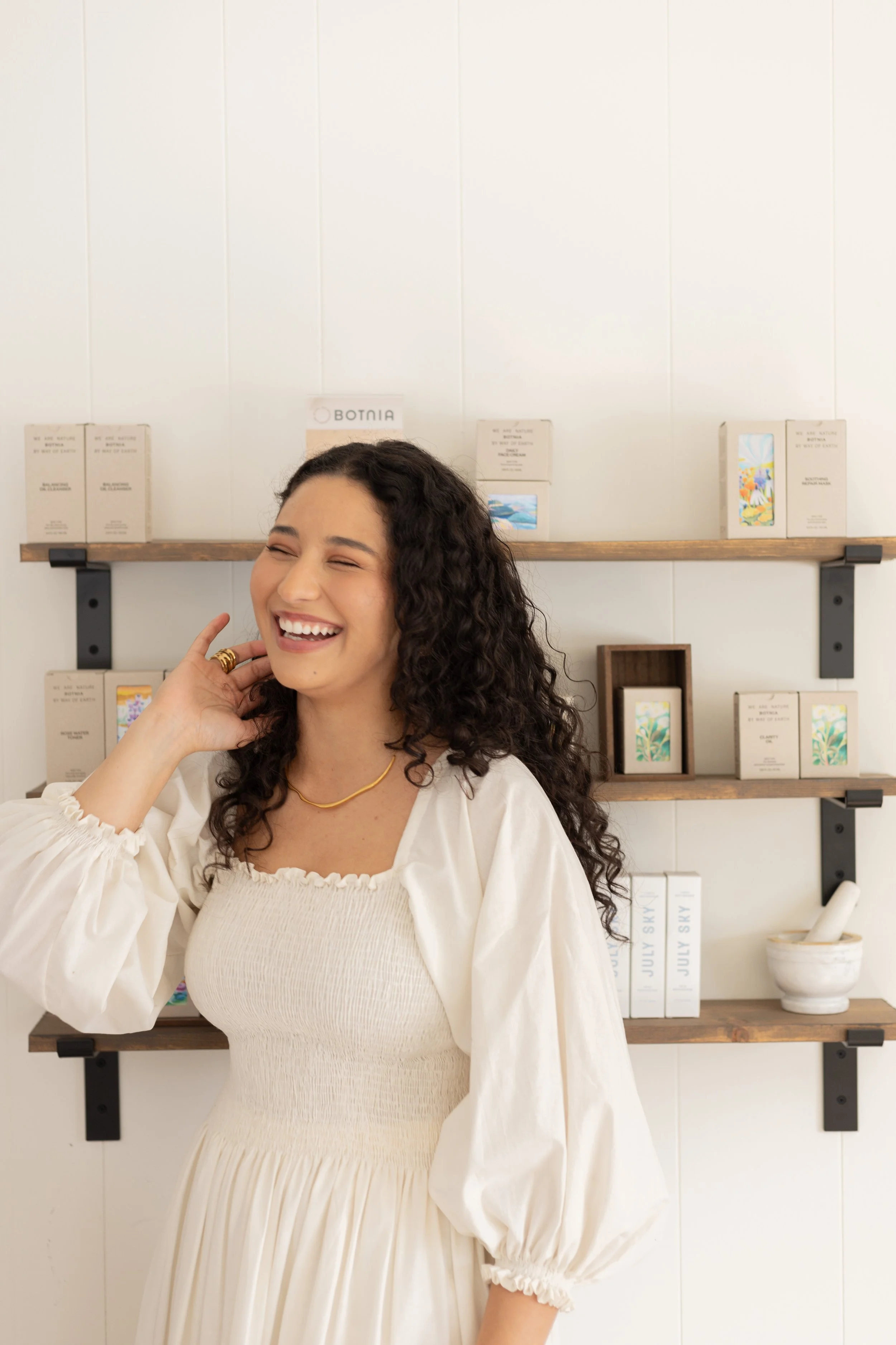 A woman with curly hair smiling and laughing, wearing a cream-colored dress with puffed sleeves, standing in front of wall-mounted wooden shelves with products and decor.