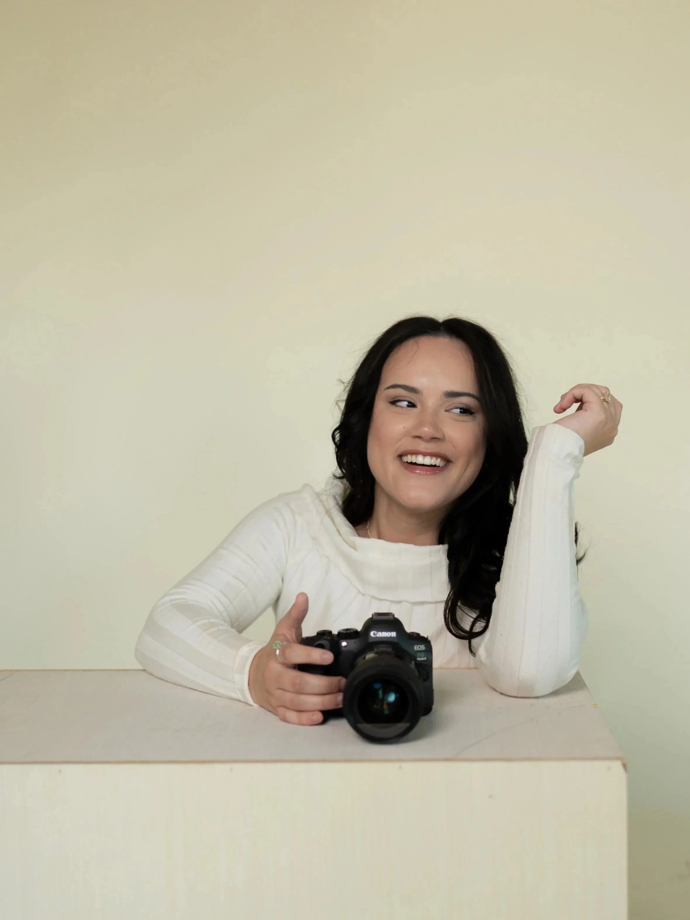 A woman with dark hair smiling and looking to her right, sitting at a table with a Canon camera in front of her, against a plain beige background.