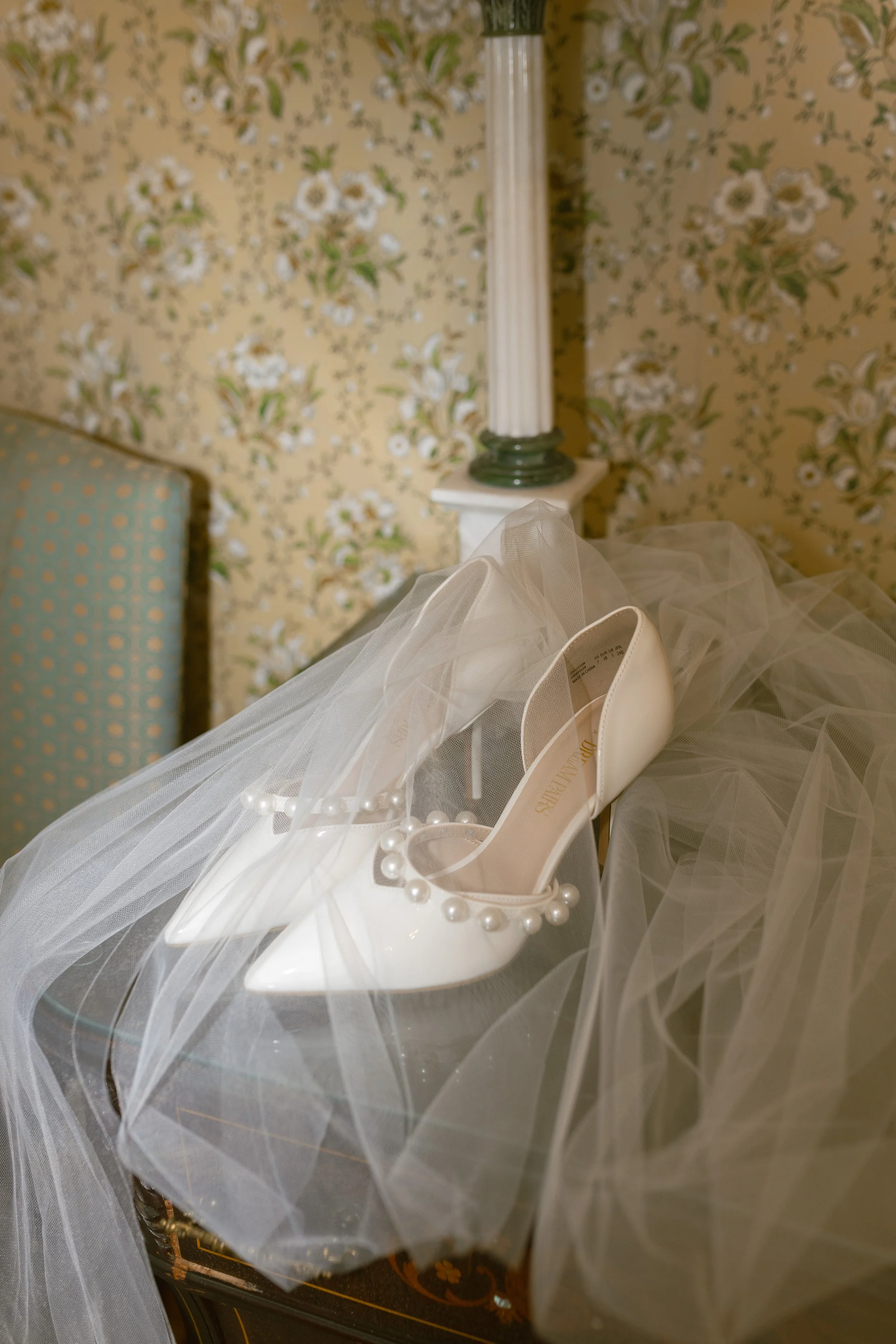 White bridal shoes with pearl embellishments on a table, surrounded by a sheer veil in a vintage-style room.