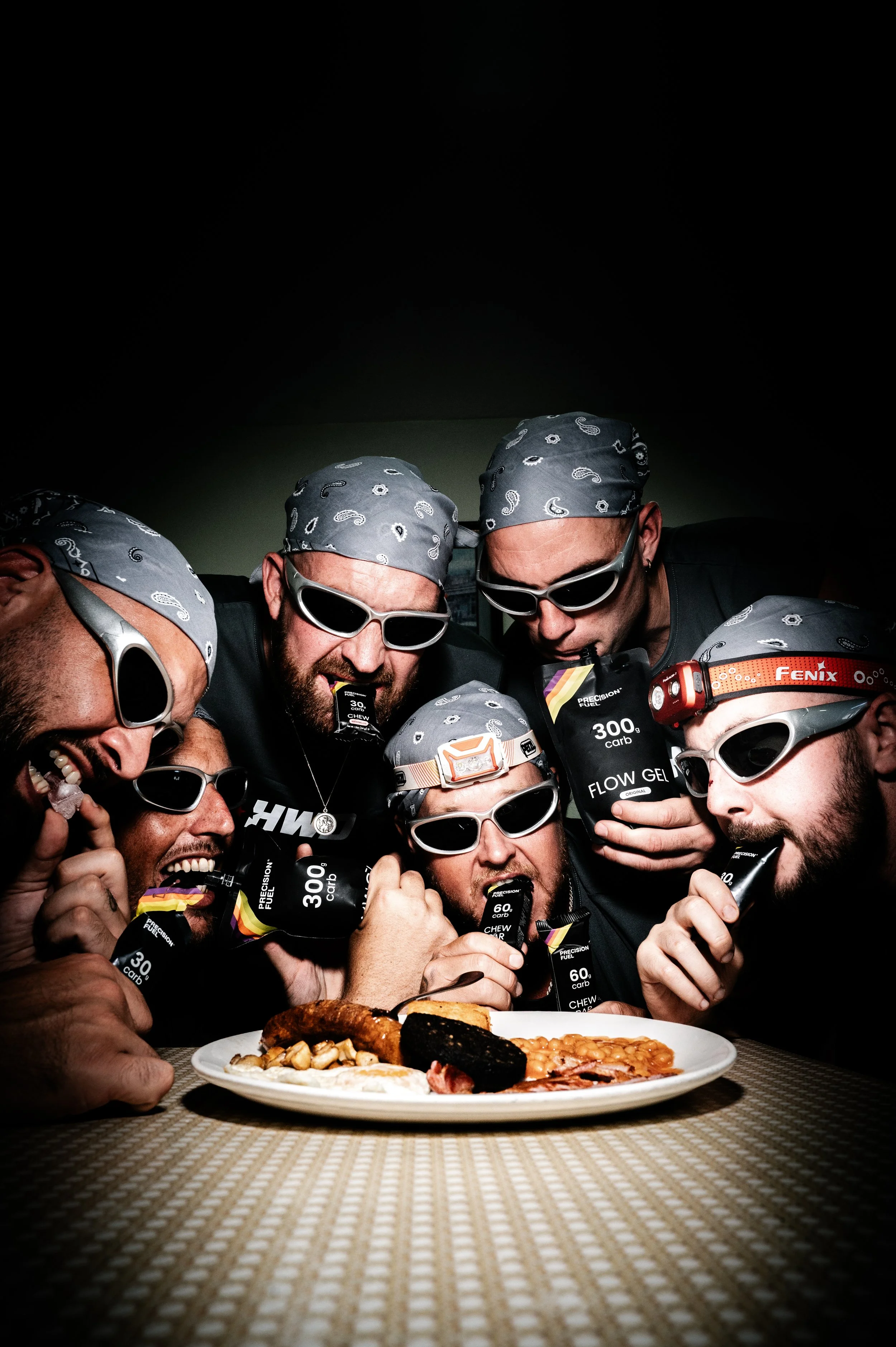 Group of people dressed in black, wearing bandanas and sunglasses, gathered around a table with a plate of food, celebrating or preparing for a meal.