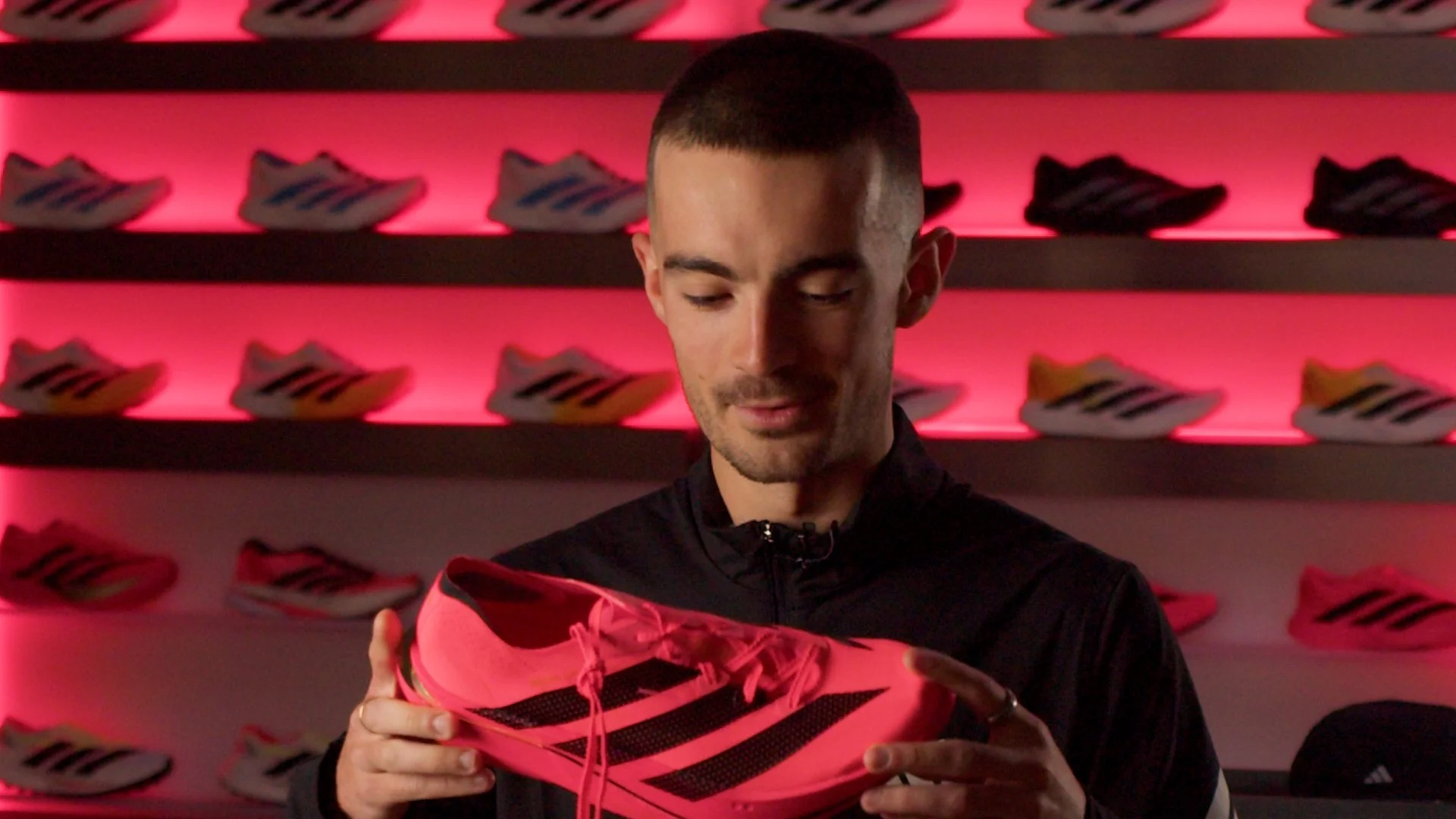 A man with short dark hair holding a bright pink and black athletic shoe in front of a wall decorated with multiple pairs of similar shoes on shelves, with pink lighting.