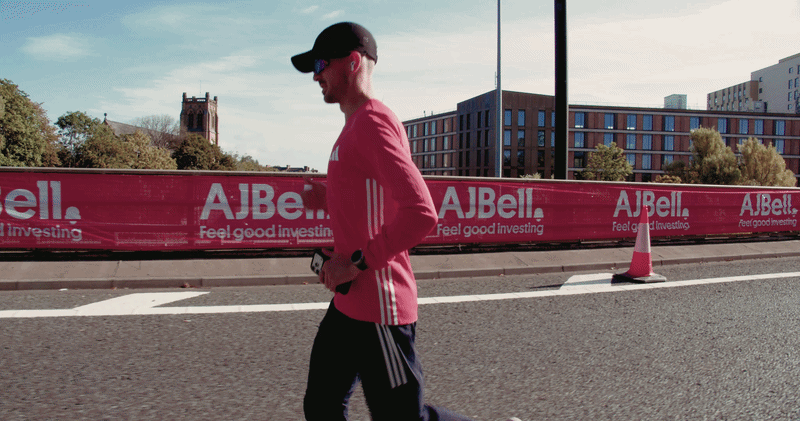 Cole Gibbens running on a city street during daytime, wearing a pink adidas top, black cap, sunglasses, and black adidas pants with white stripes. There's a pink Great north Run barricade with advertising and a traffic cone in the background.