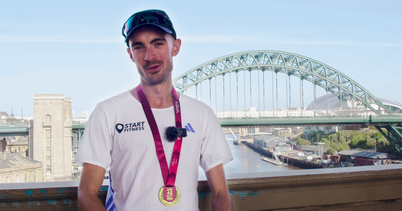 cole gibbens wearing a white start fitness and adidas shirt and a great north run medal around his neck, standing outdoors with a river and a the tyne bridge in the background.
