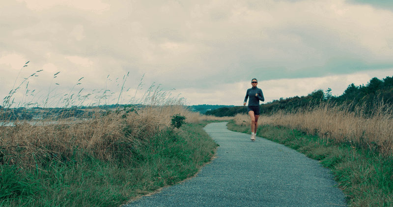 nike athlete cari hughes running on a paved trail through a grassy countryside landscape with trees in the background and a cloudy sky.