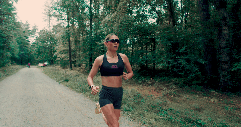 cari hughes running on a gravel road through a forest, wearing sunglasses, a sports bra, and shorts.