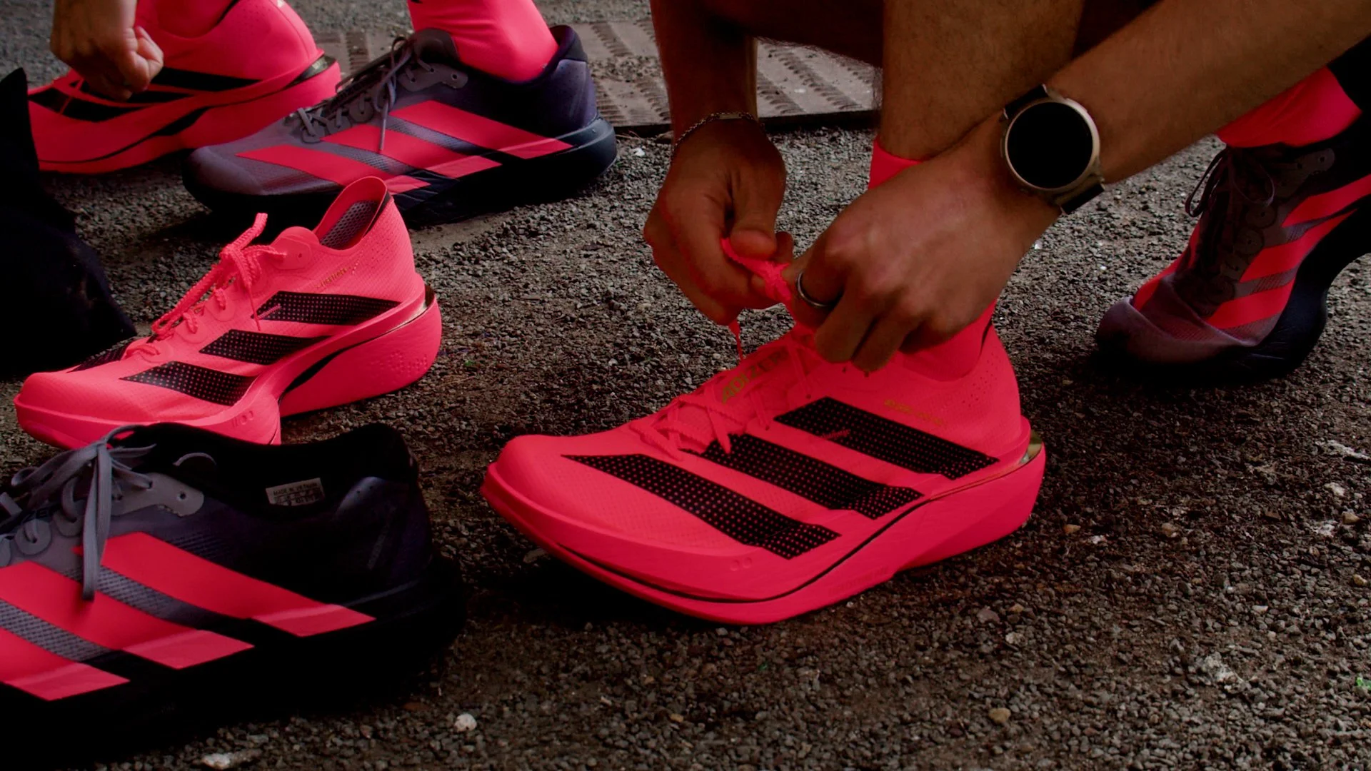 Person tying bright pink Adidas running shoes next to other pink and black athletic shoes on gravel ground.